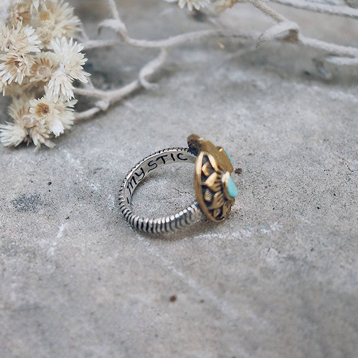 The Natural Mystic Ring - Turquoise by bahgsu jewels is an adjustable sterling silver ring engraved with MYSTIC and features a gold and turquoise top. Shown on textured stone near dried white flowers, it adds unique flair to any collection.