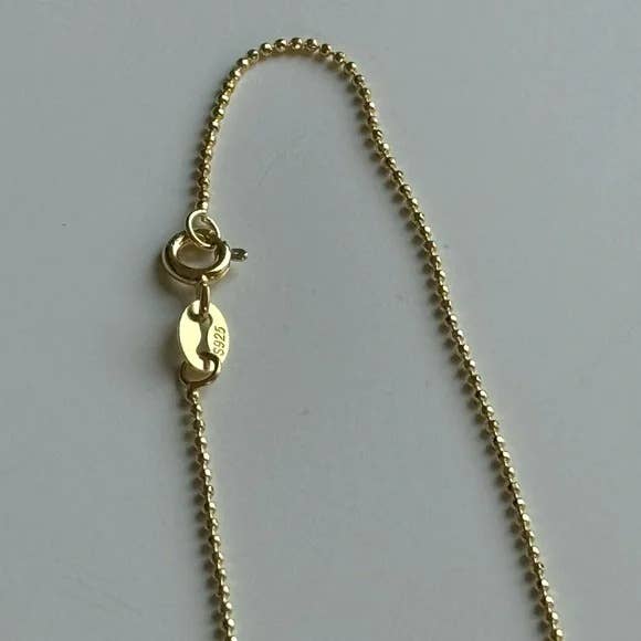 A close-up of the Tramps+Thieves Scarab Necklace, featuring gold beads, a round clasp, and a tag marked 925 for sterling silver under gold plating, set against a light background.
