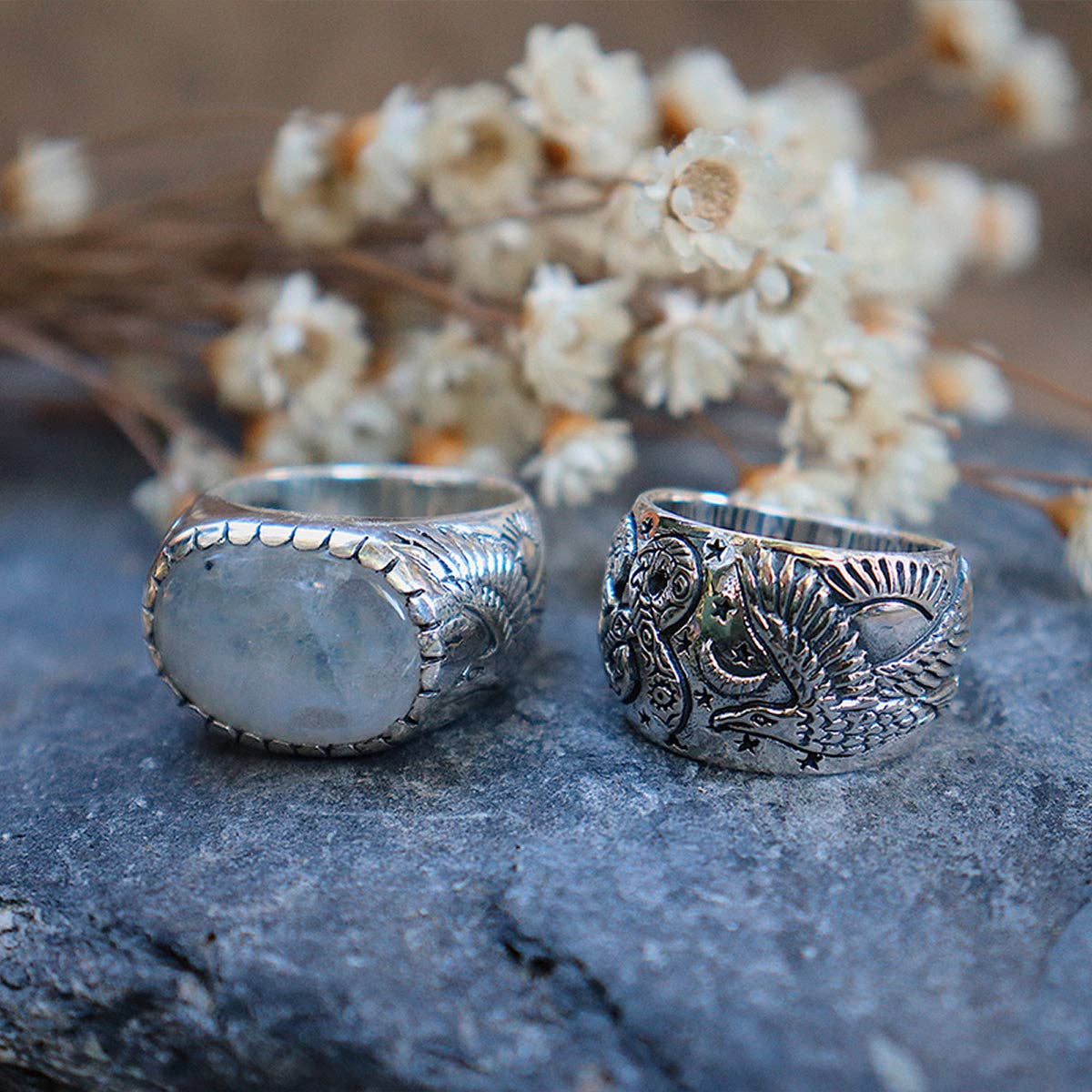 Two ornate bahgsu jewels rings rest on stone. One is the Rainbow Serpent Ring from the Xavier Rudd Collab, showcasing an oval moonstone, while the other displays intricate engravings. Dried flowers blur gently in the background.