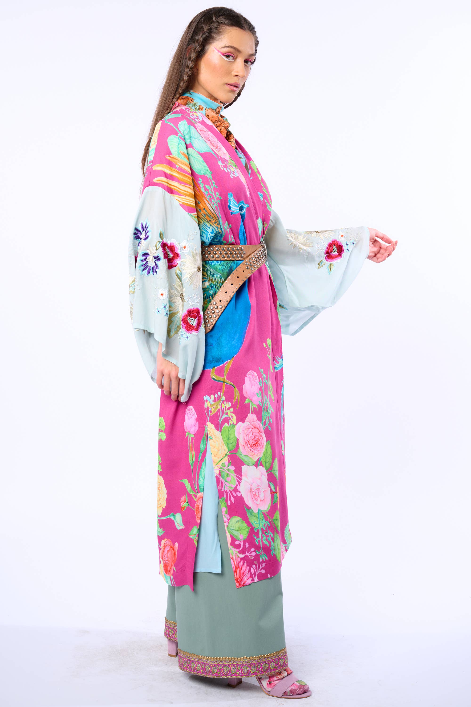 A woman stands sideways in the Aratta Marie Antoinette Kimono, featuring bright pink, blue, and green floral embroidered sleeves, a decorated obi belt, and matching embroidered shoes against a plain white background.