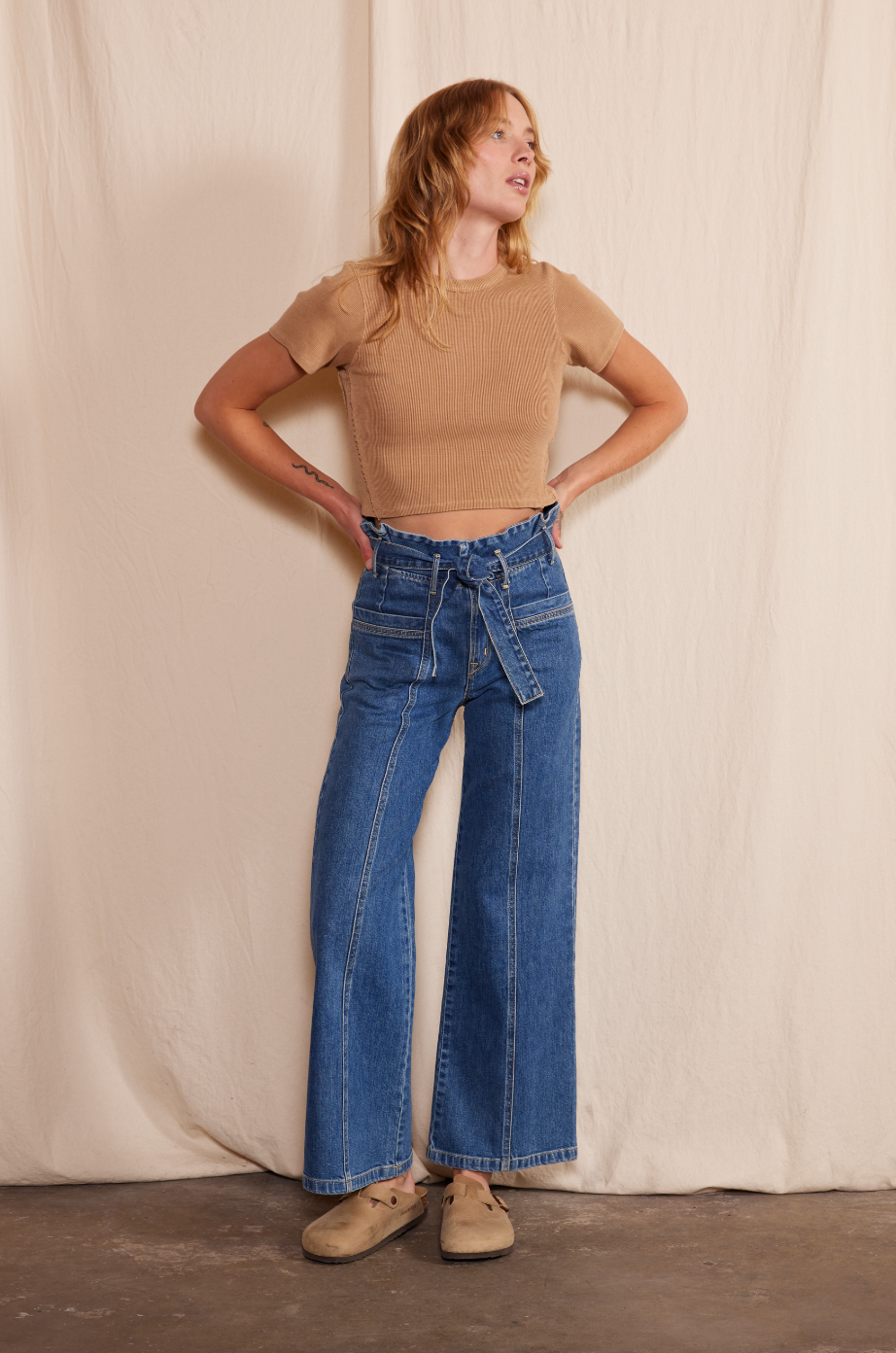 A woman stands before a beige fabric backdrop, wearing a short-sleeve beige top and Porter Blue Apparels PENNY PAPERBAG WIDE LEG IN LAGUNA WASH jeans, crafted from organic cotton denim by Candiani Mills, paired with tan slip-on shoes.