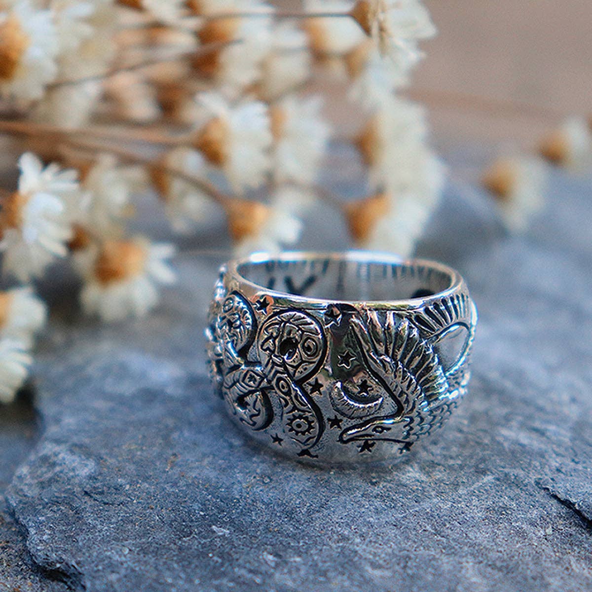 The Rainbow Serpent Ring by bahgsu jewels, a striking sterling silver piece from the Xavier Rudd Collab, features intricate serpent and celestial engravings, shown resting on gray stone with dried white flowers in the background.