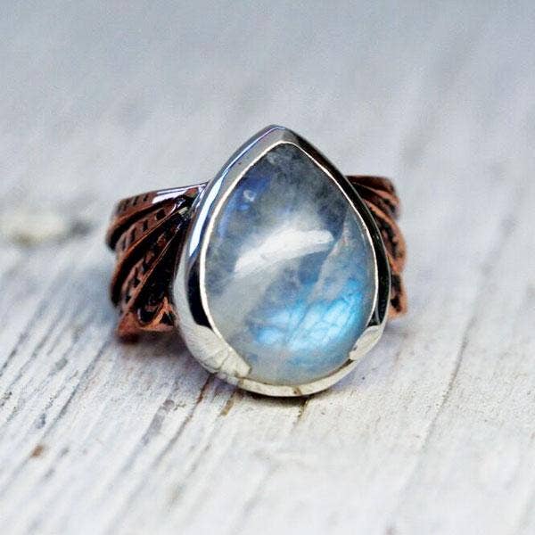 The Winged Ring by bahgsu jewels features a teardrop moonstone in a silver bezel with a copper band and leaf details. This adjustable brass ring is shown resting on light wood.