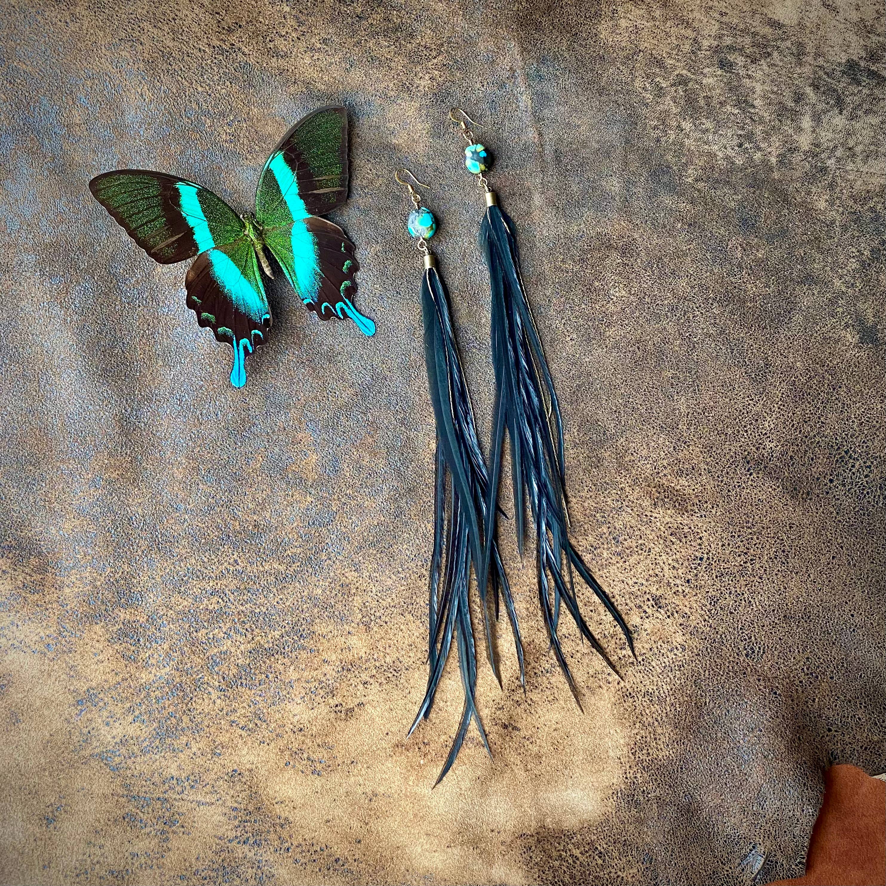 A blue and black butterfly rests beside ASTALIs Feather & Turquoise Statement Earrings—made with antique brass, turquoise studs, and sustainable coque feathers—displayed on a textured brown surface.