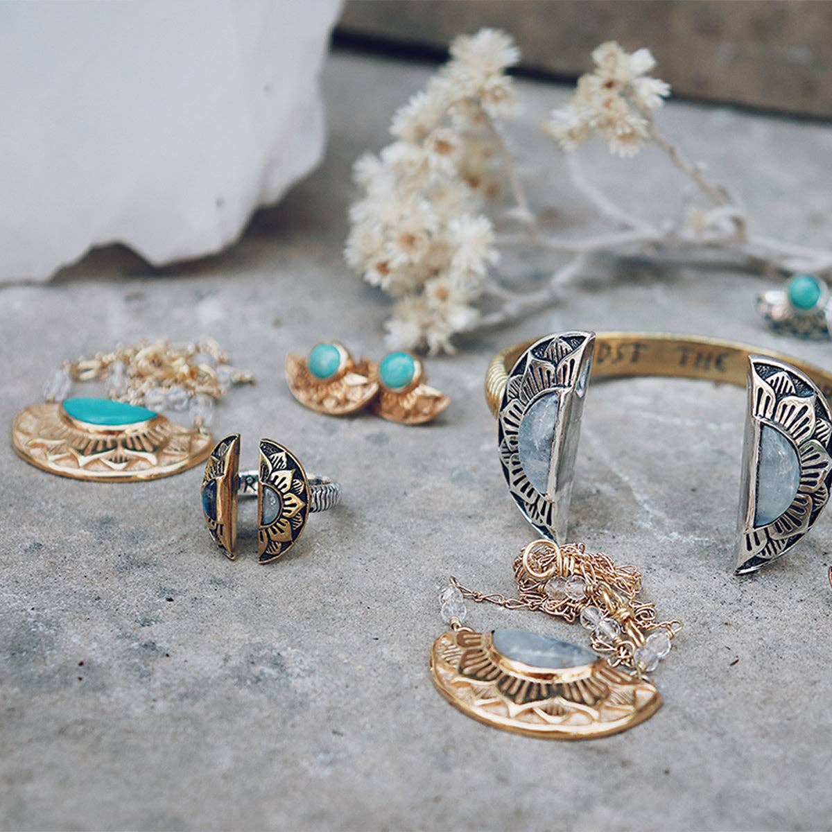 Bahgsu Jewels Natural Mystic Ring—Moonstone, along with gold and sterling silver rings, necklaces, earrings, and an adjustable two-toned ring, are arranged on stone with dried flowers in the background for a bohemian display.