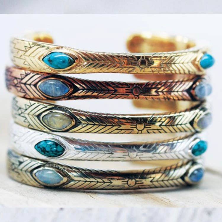 A stack of bahgsu jewels Medicine Wheel Cuffs in gold, copper, and Indian silver tones features engraved designs with oval turquoise or blue stone inlays, beautifully displayed on a light surface.