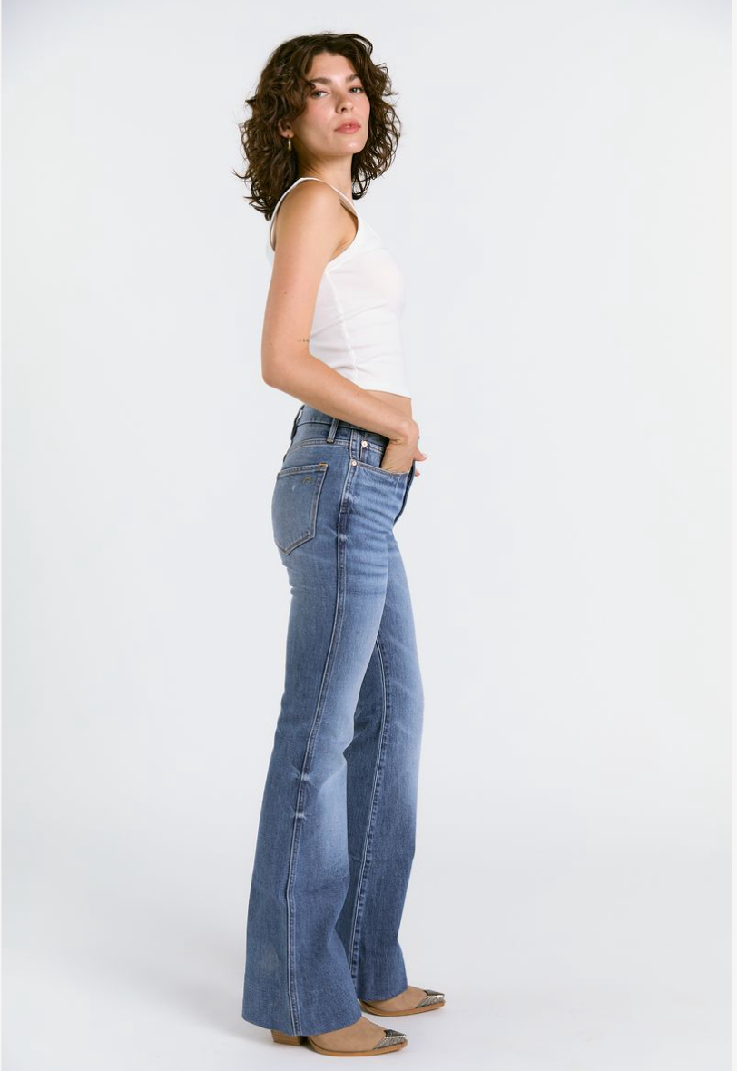 A woman with curly hair stands sideways against a white background, wearing the Porter Blue Apparel WANDERER 5 POCKET FLARE IN MABEL WASH jeans, a white sleeveless top, and beige shoes. She poses confidently with one hand in her pocket.