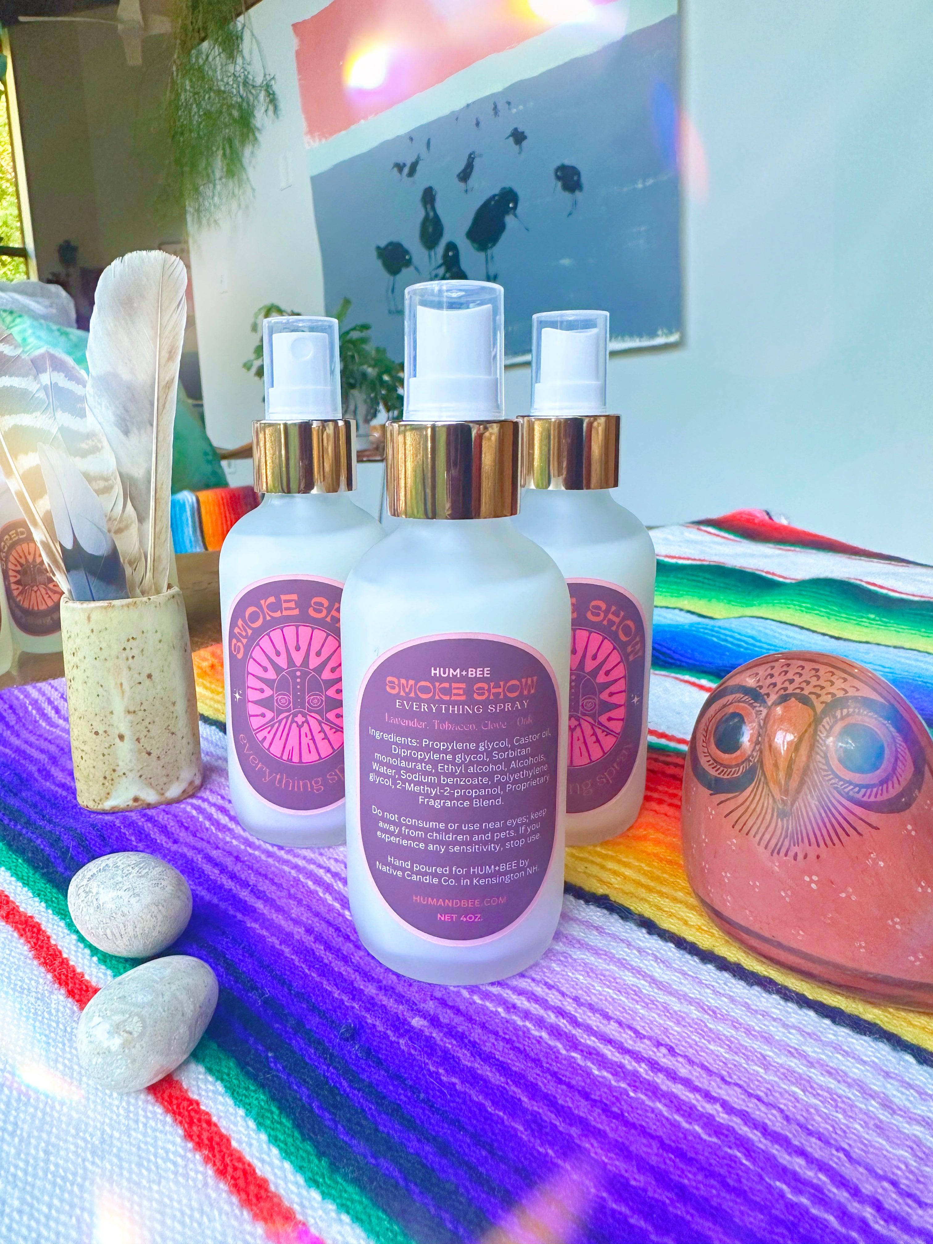 Four bottles of HUM+BEE Smoke Show Everything Spray rest on a colorful striped cloth beside a ceramic owl, two white stones, and feathers. This hand-poured spray brings a smoky, inviting fragrance—perfect for any room. Modern art and greenery complete the scene.