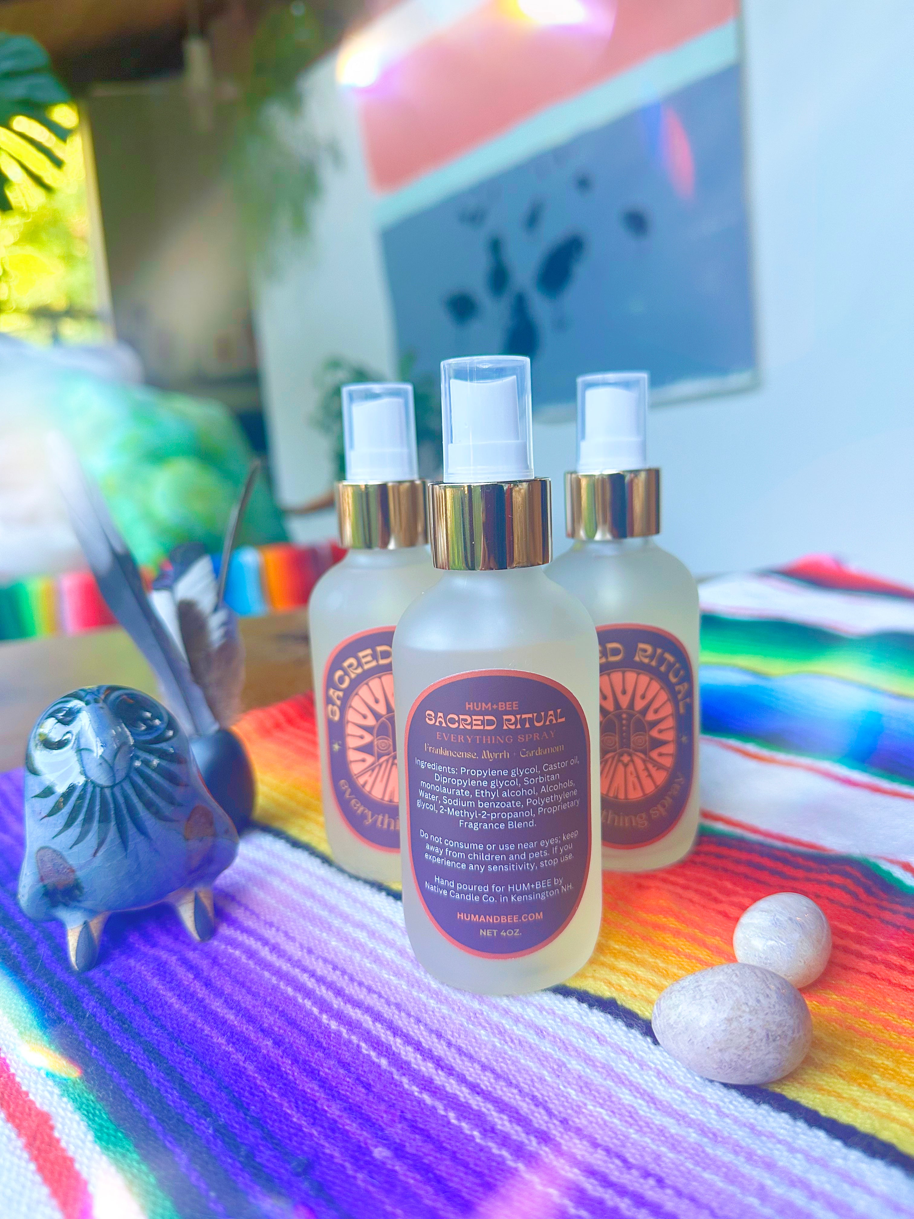 Three bottles of HUM+BEE Sacred Ritual Everything Spray rest on a colorful striped cloth beside a bird figurine and two stones, with vibrant decor and greenery in the background.