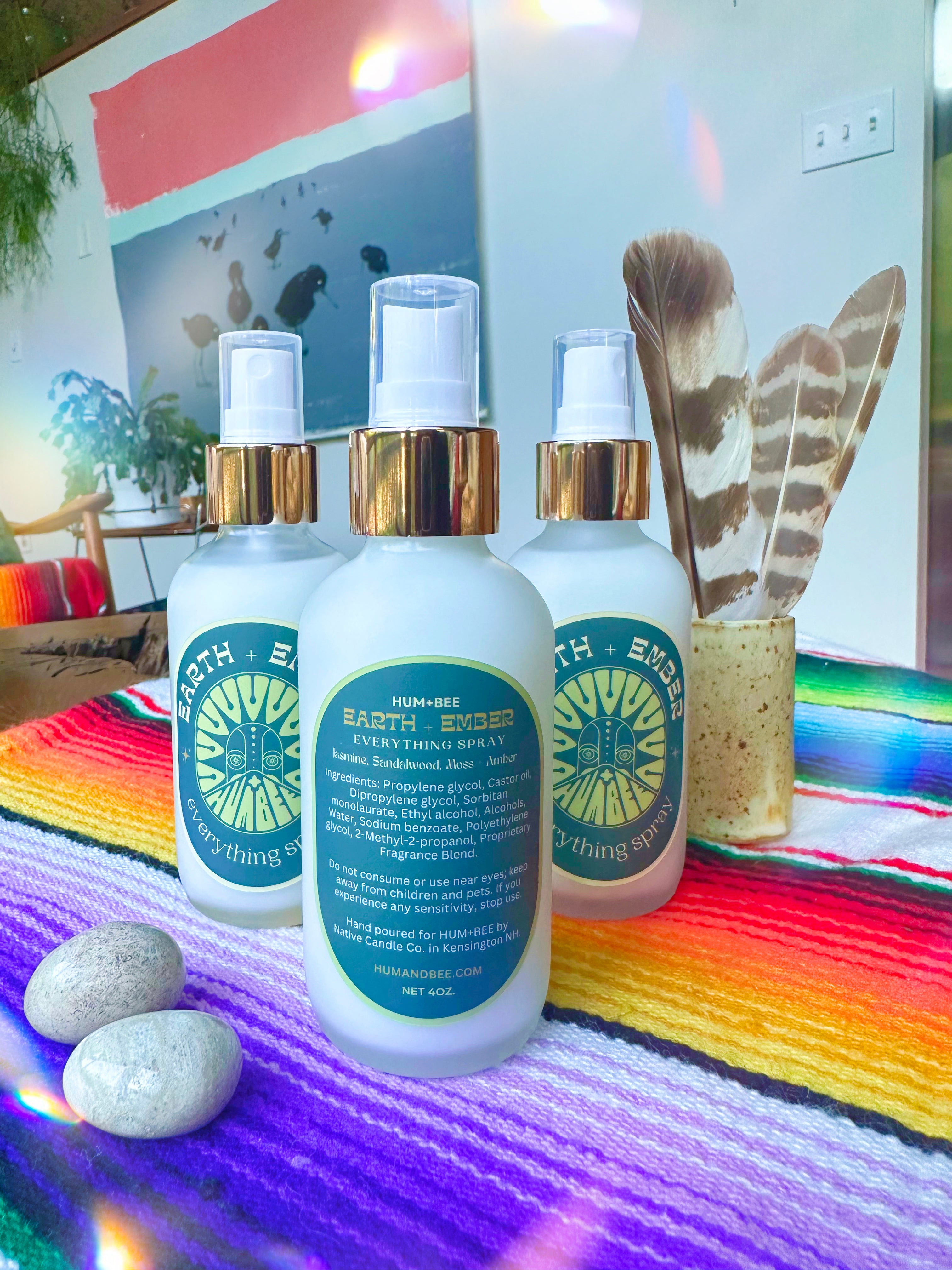 Three bottles of HUM+BEE Earth + Ember Everything Spray, a vibrant cedarwood amber home fragrance, are displayed on a colorful striped cloth with rocks and feathers nearby, set against a striking wall mural.