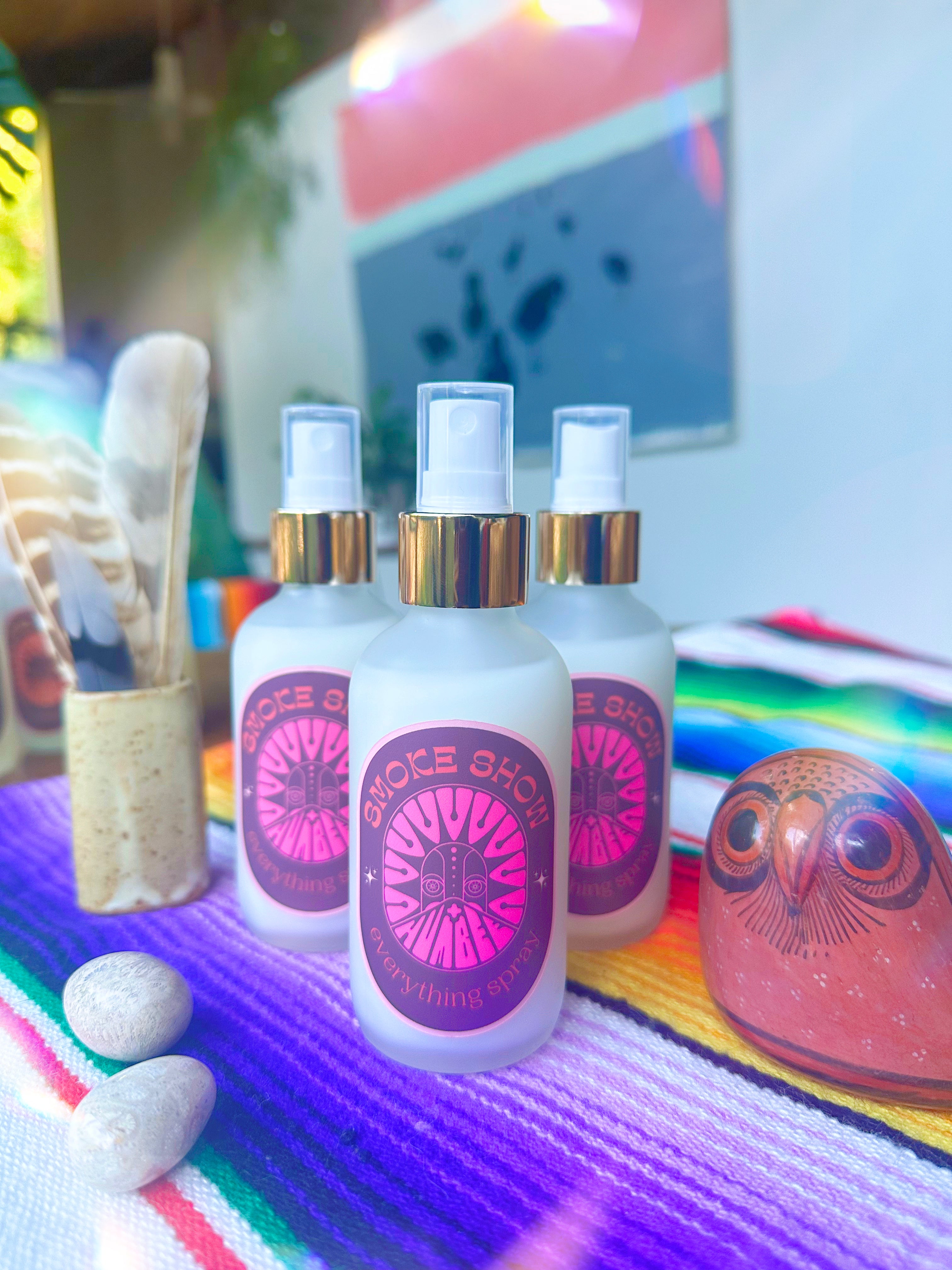 Three bottles of HUM+BEE's Smoke Show Everything Spray rest on a vibrant striped cloth near a carved owl, stones, and feathers—this hand-poured fragrance is ideal for anyone who loves smoky scents.