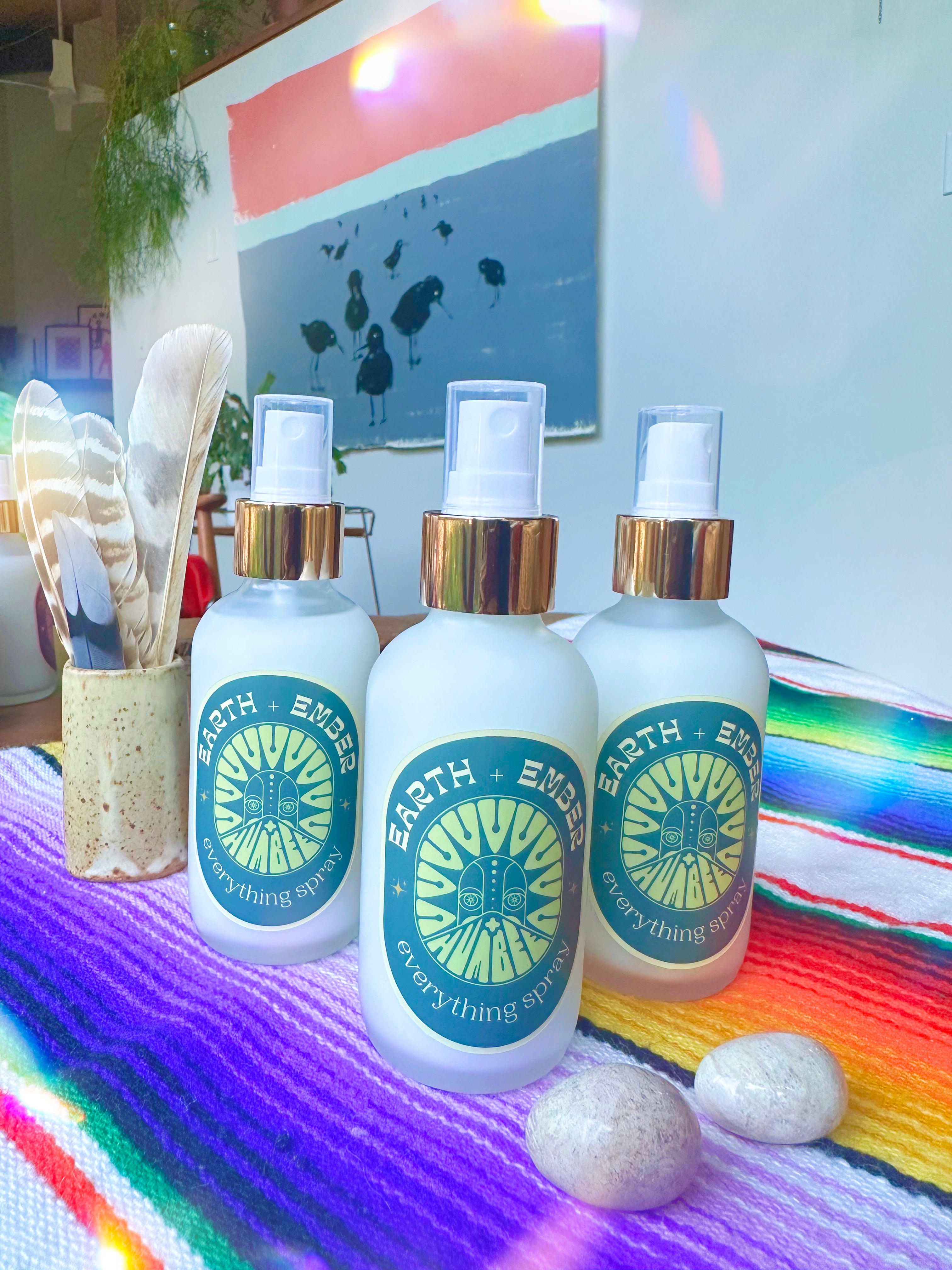 Three bottles of HUM+BEE’s Earth + Ember Everything Spray rest on a vibrant striped cloth with stones, feathers, and a large bird painting in the background—an inviting scene highlighting this cedarwood amber home fragrance.