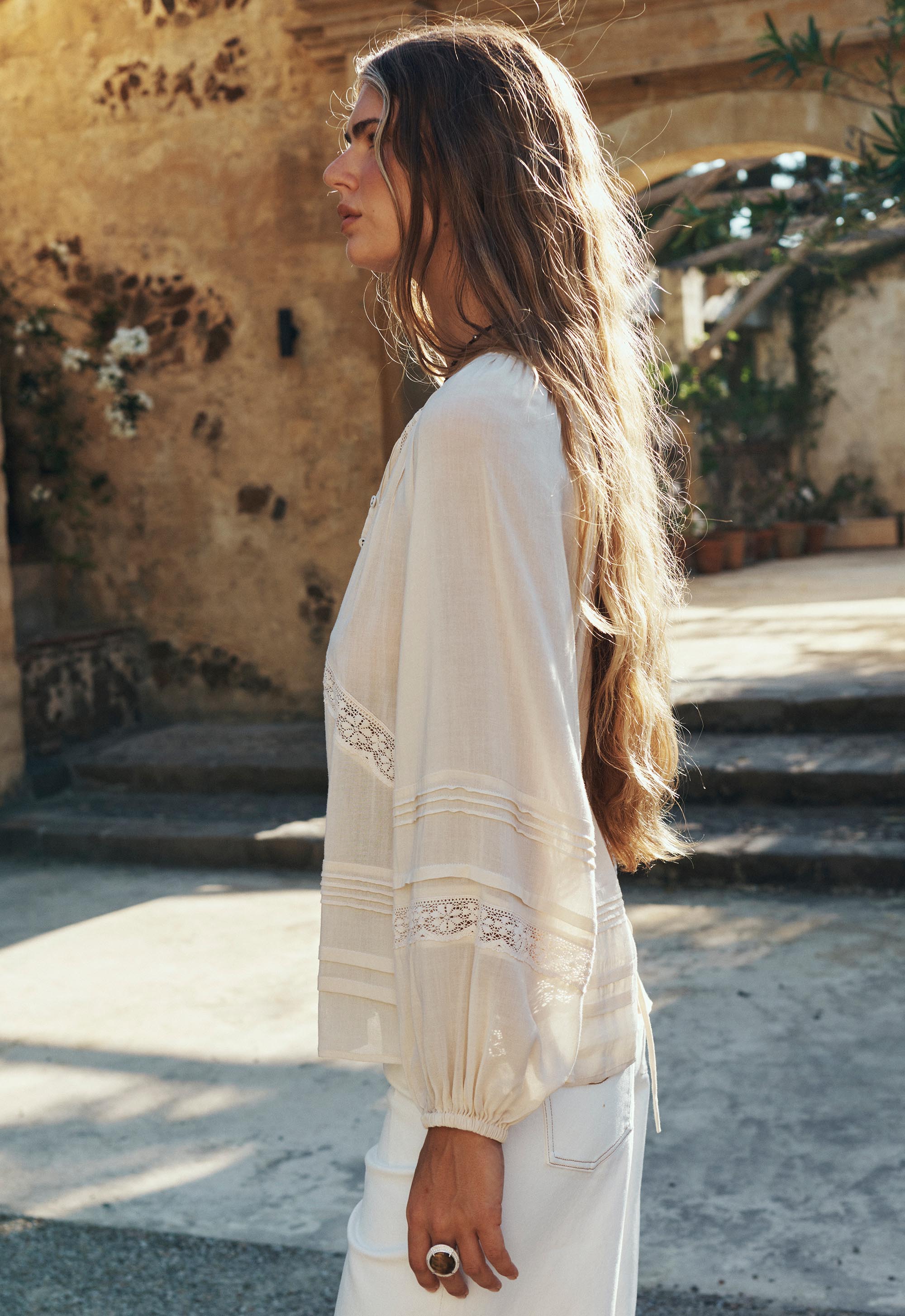 Woman wearing Hera Lace Blouse- Macadamia with delicate lace details and flowing sleeves in sunlit rustic courtyard