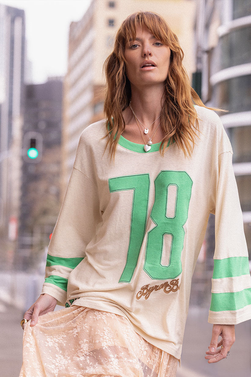 Woman wearing a Homecoming Long Sleeve Biker Tee with green number 78 and green striped sleeves in urban setting
