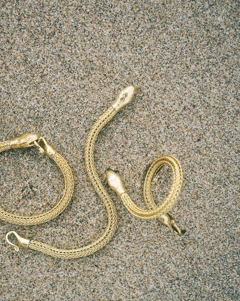 Serpent Bracelet in gold tone with detailed scales, shaped like a coiled snake, lying on textured sand surface