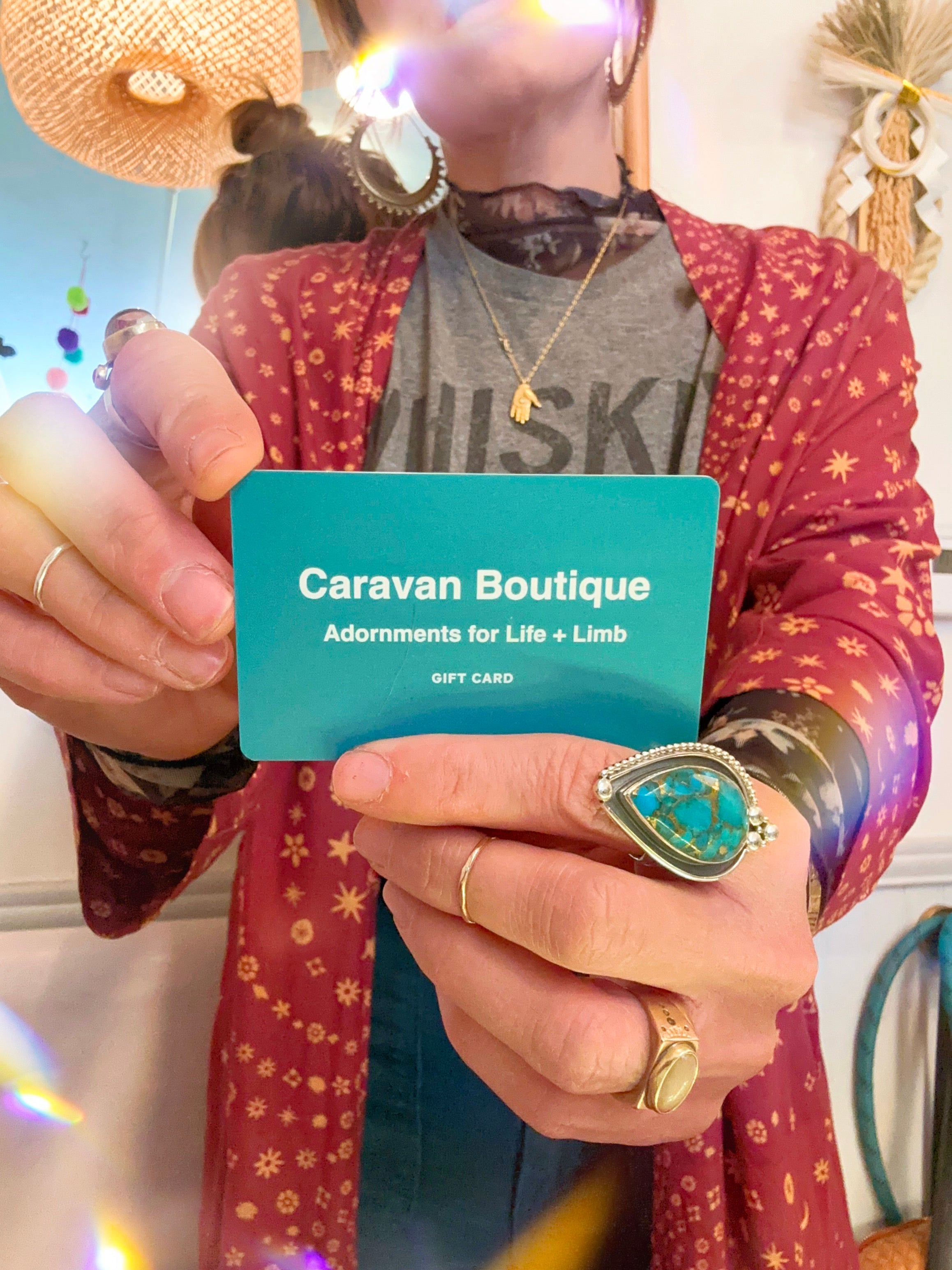 A person in a red patterned cardigan and rings holds a turquoise Caravan Boutique Gift Card by Caravan, with “Adornments for Life + Limb” visible—ideal for gifting or your next Styling Room session.