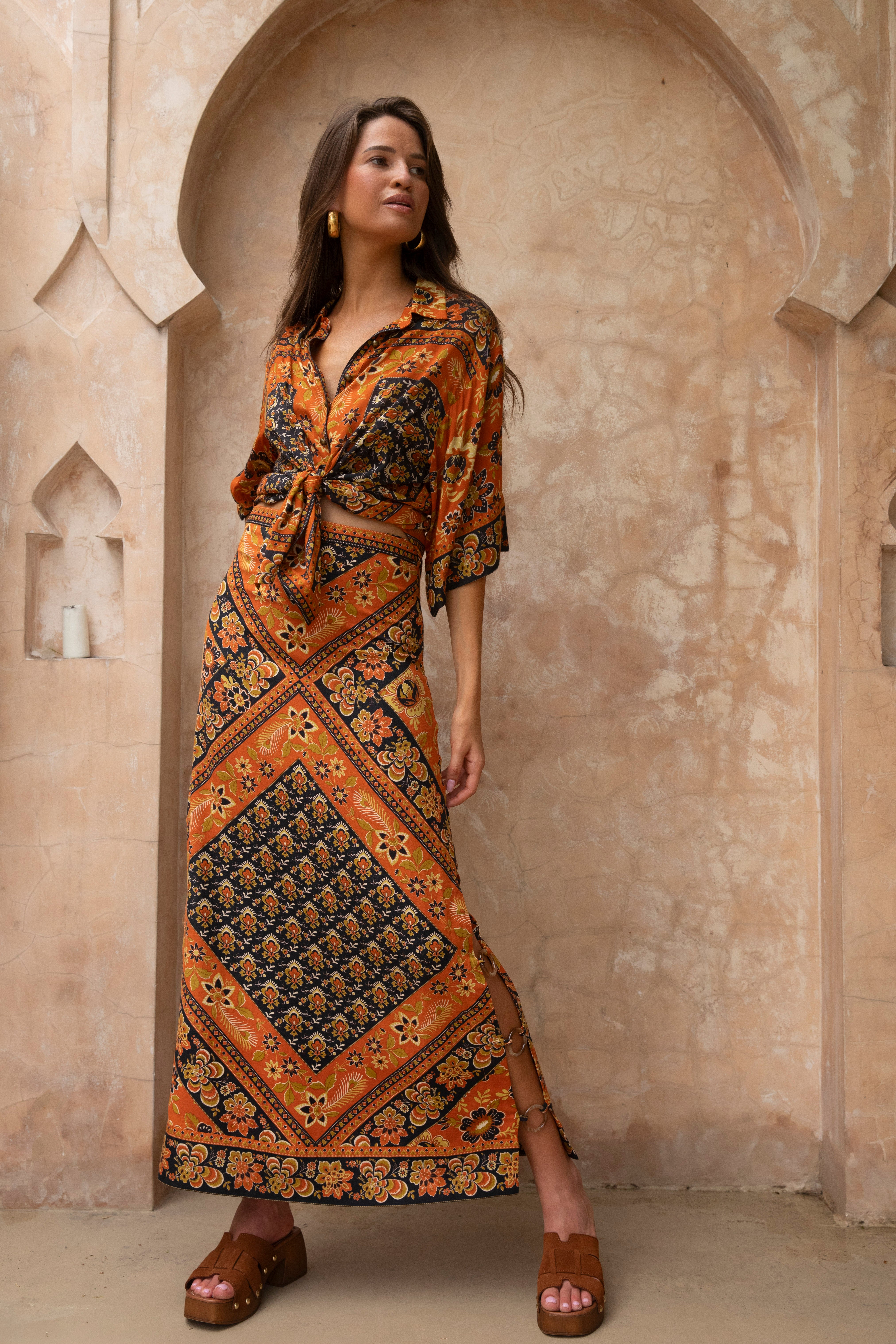 Woman wearing a patterned maxi skirt and tied front Lois Shirt - Saffron posing against textured arch wall