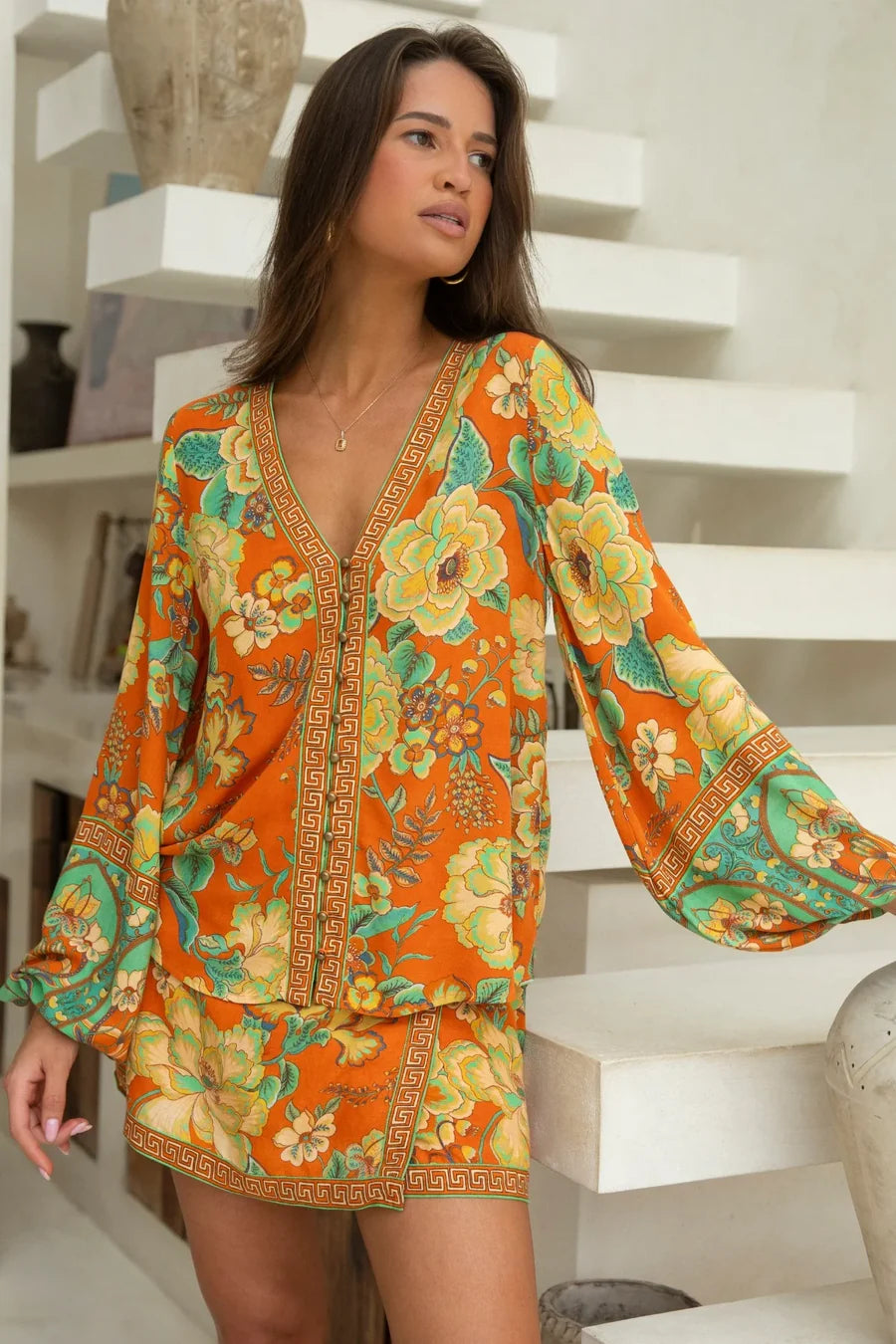 A woman stands indoors by white shelves, gazing sideways with one hand on a surface, wearing the Nine Lives Bazaar Maddison Blouse - Tango featuring vibrant orange florals and poet sleeves.