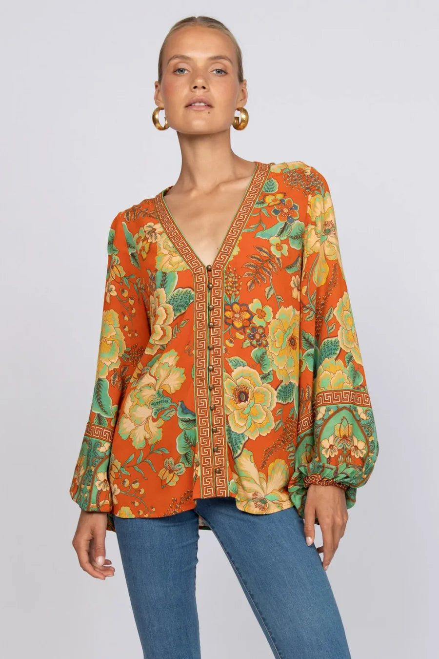 A woman wears the Nine Lives Bazaar Maddison Blouse - Tango, a vibrant viscose crepe top with poet sleeves, a yellow floral pattern, and Greek key trim, styled with blue jeans against a neutral backdrop.