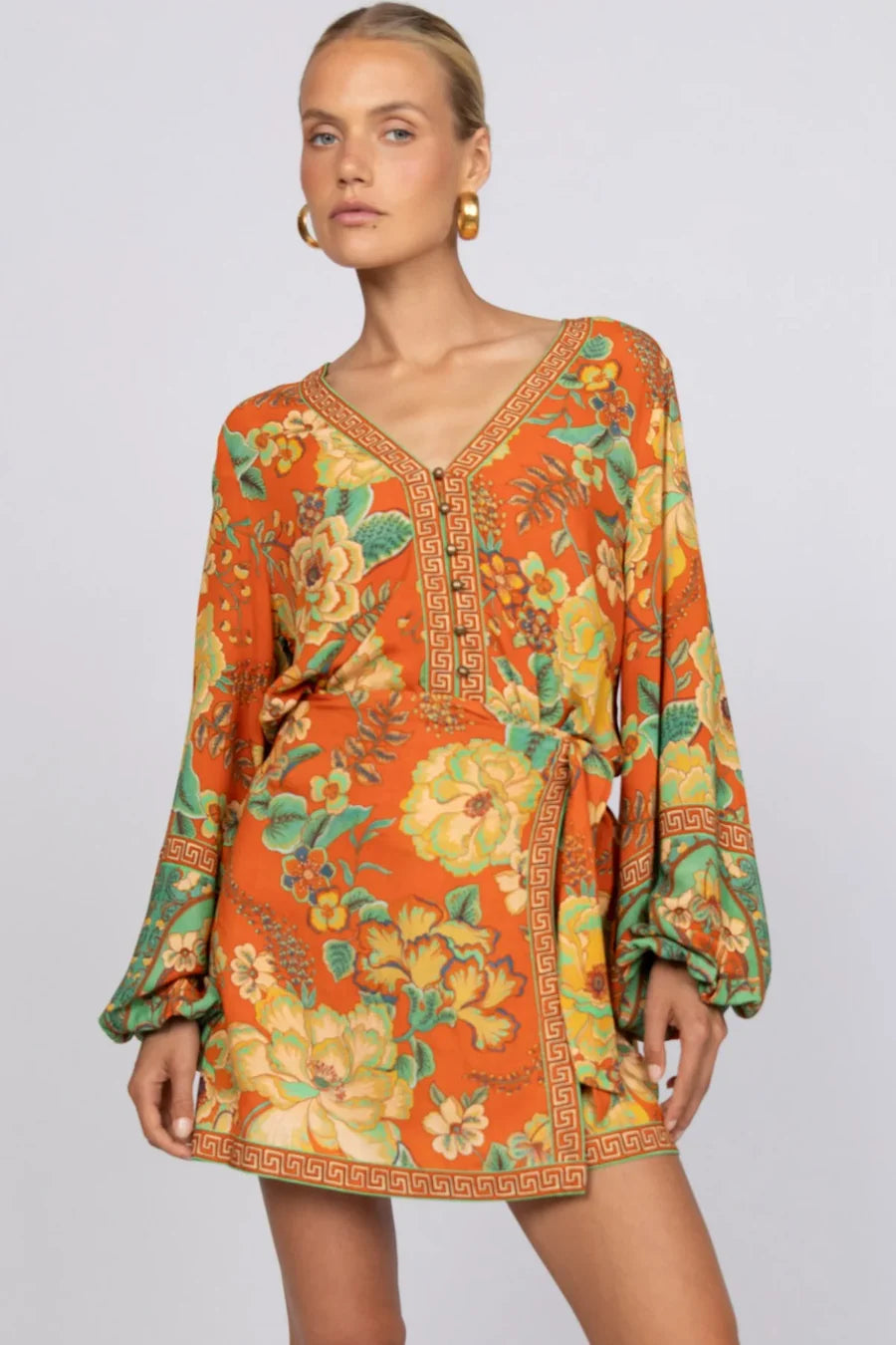 A woman models the Maddison Blouse - Tango by Nine Lives Bazaar, featuring a vibrant orange floral print with poet sleeves and a V-neckline with a Greek key border, paired with gold hoop earrings and hair pulled back against a light background.