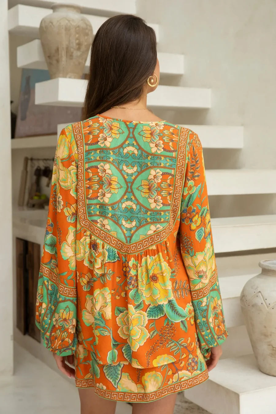 A woman with long brown hair and gold hoop earrings, wearing the vibrant Maddison Blouse - Tango by Nine Lives Bazaar, stands with her back to the camera in a stylish, light-colored indoor space.