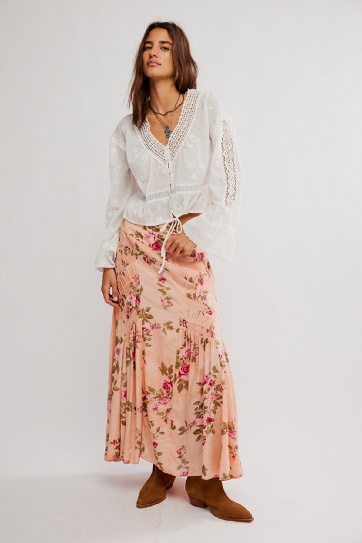A woman stands against a plain backdrop wearing a white long-sleeve embroidered blouse, brown boots, layered necklaces, and the Free People Rowan Maxi Skirt featuring a rose floral print and A-line silhouette. She looks slightly to the side, relaxed.
