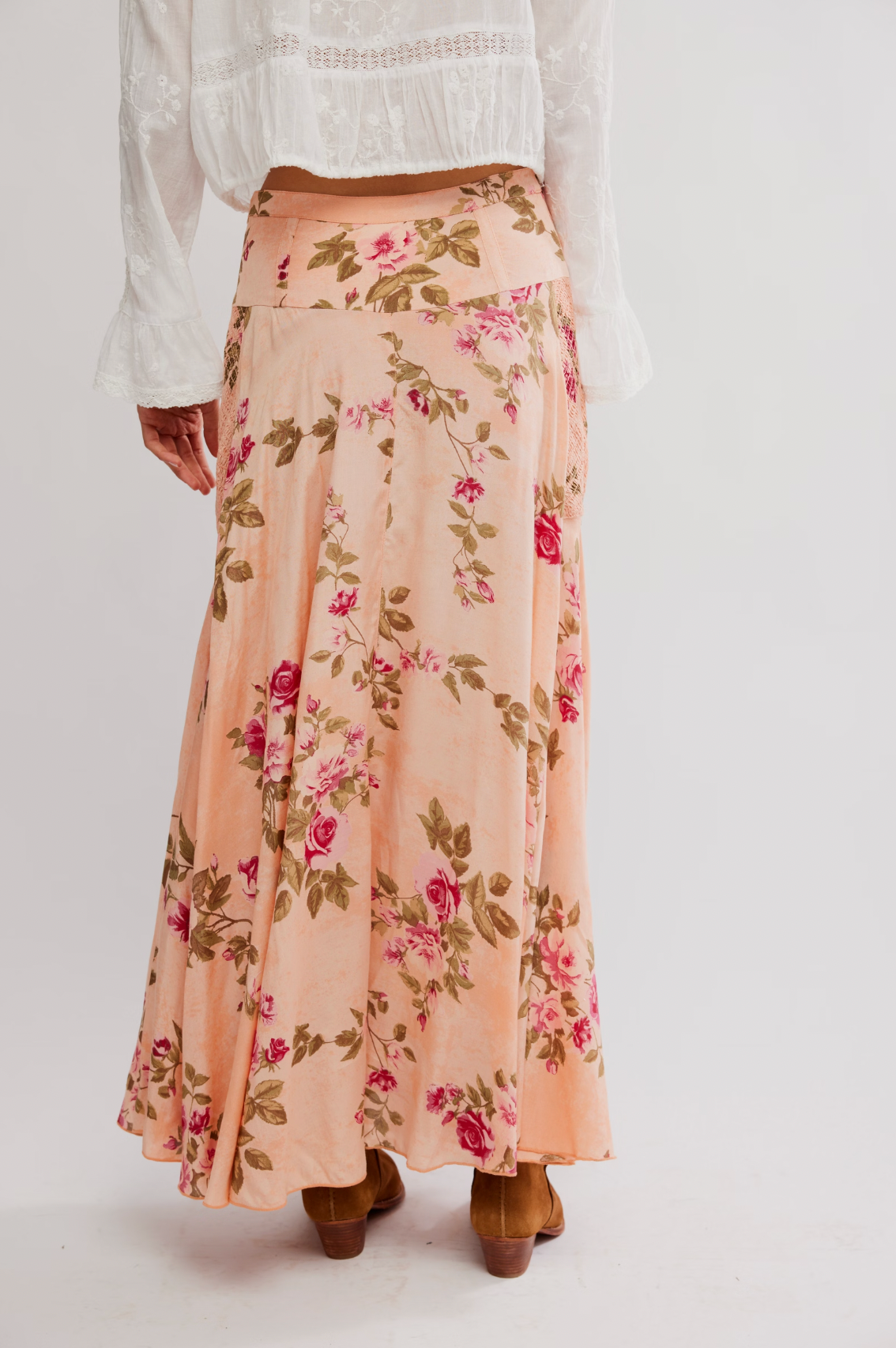 A person wearing the Rowan Maxi Skirt by Free People, featuring a peach rose floral print, paired with a white lace-detailed blouse and brown heeled boots, seen from the back against a plain background.
