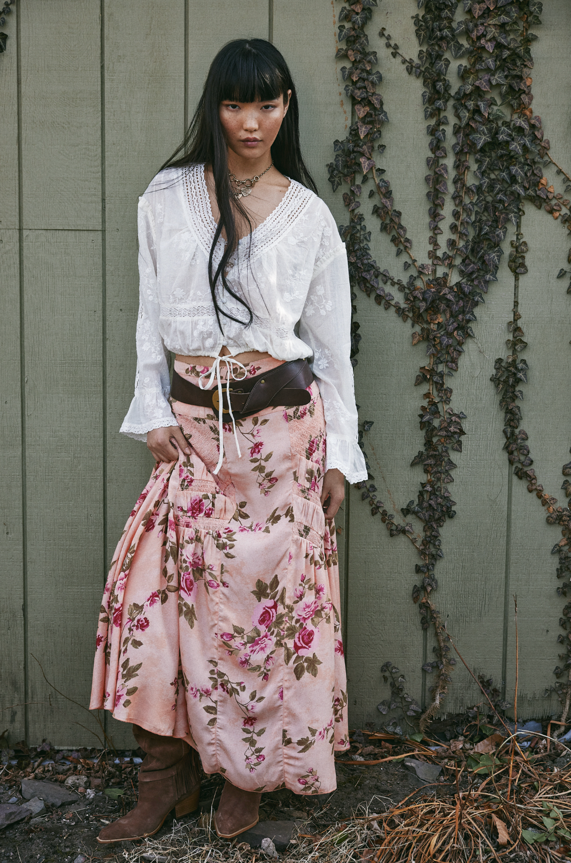A woman with long dark hair and bangs stands against a wooden wall with ivy, wearing the Free People Rowan Maxi Skirt in a rose floral print, paired with a white blouse, brown boots, layered necklaces, and a neutral expression.