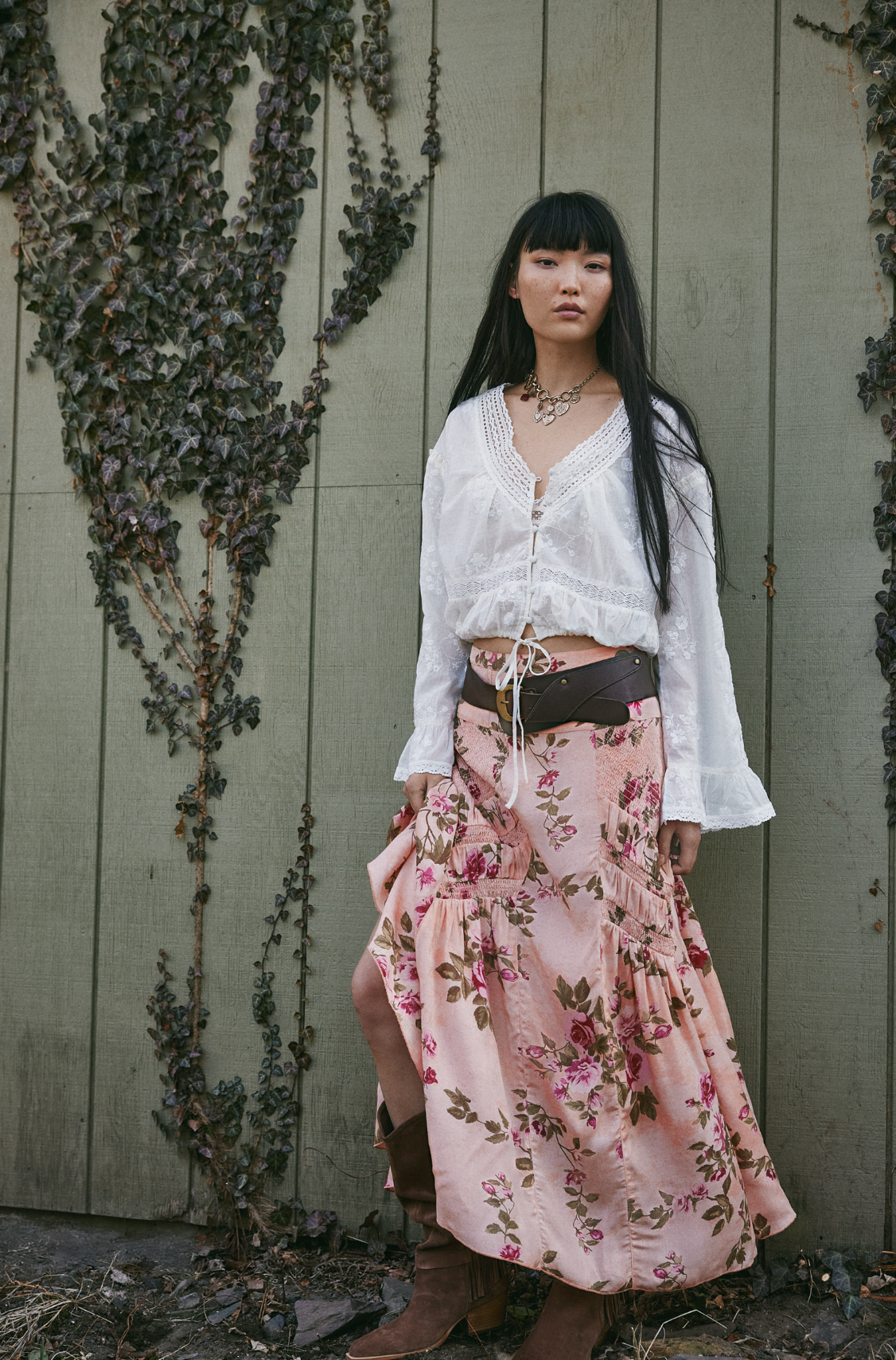 A woman with long black hair and a neutral expression stands against a vine-covered green wall, wearing the Free People Rowan Maxi Skirt, a white long-sleeve blouse, brown belt, and brown boots.