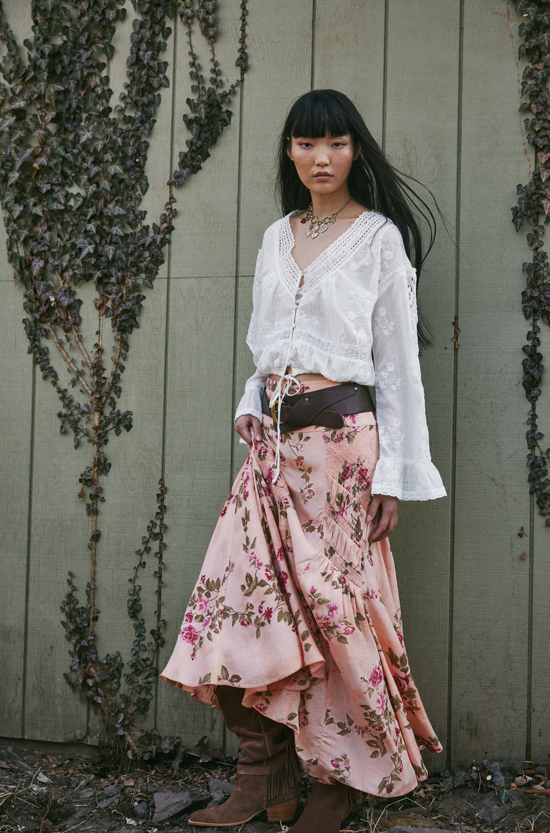 A woman with long dark hair and bangs stands by a vine-covered wall, wearing a white lace blouse and the Free People Rowan Maxi Skirt, featuring a pink rose floral print. She accessorizes with a brown belt and boots, looking serious.