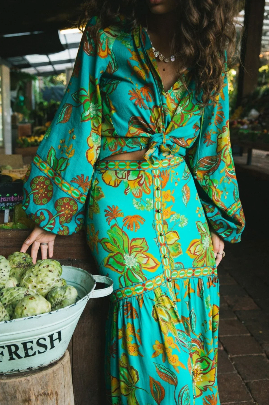 A woman in a vibrant Peacock Matilda Maxi Skirt by Nine Lives Bazaar stands by a white bucket labeled Fresh, filled with green prickly pears at an outdoor market, her wavy hair and colorful outfit showcasing bohemian style.