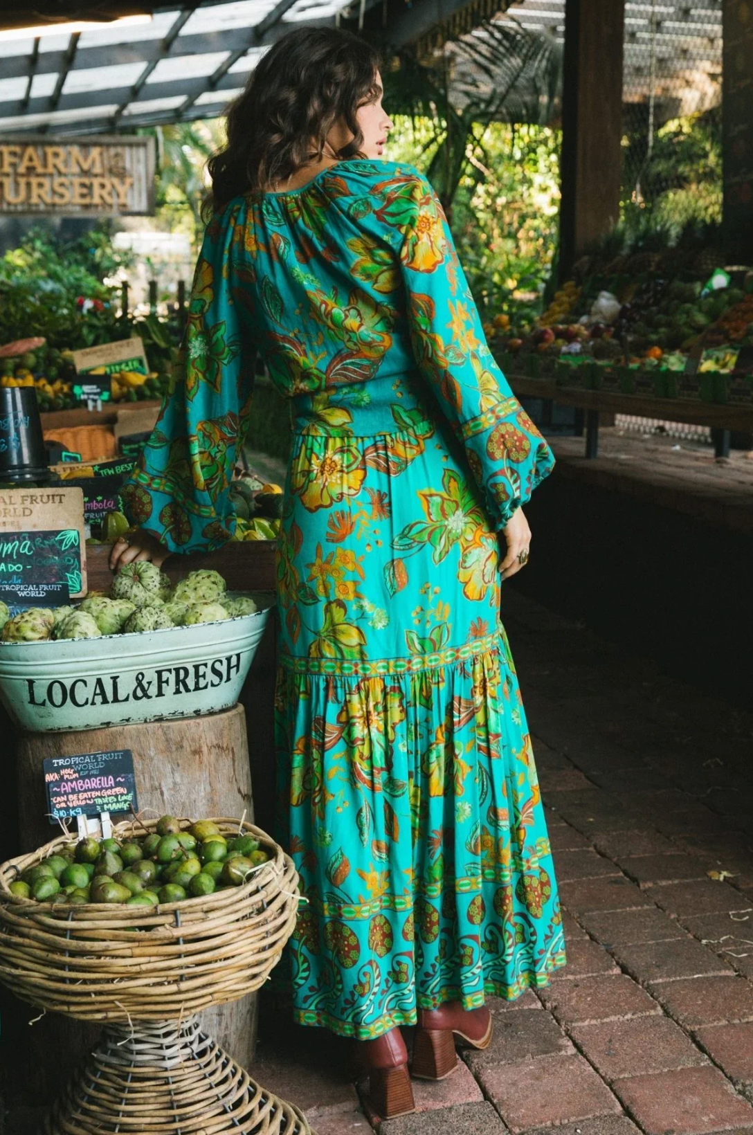 A woman in the Nine Lives Bazaar Matilda Maxi Skirt in Peacock stands facing away at an outdoor market, surrounded by baskets of fresh produce and a “Local & Fresh” sign.
