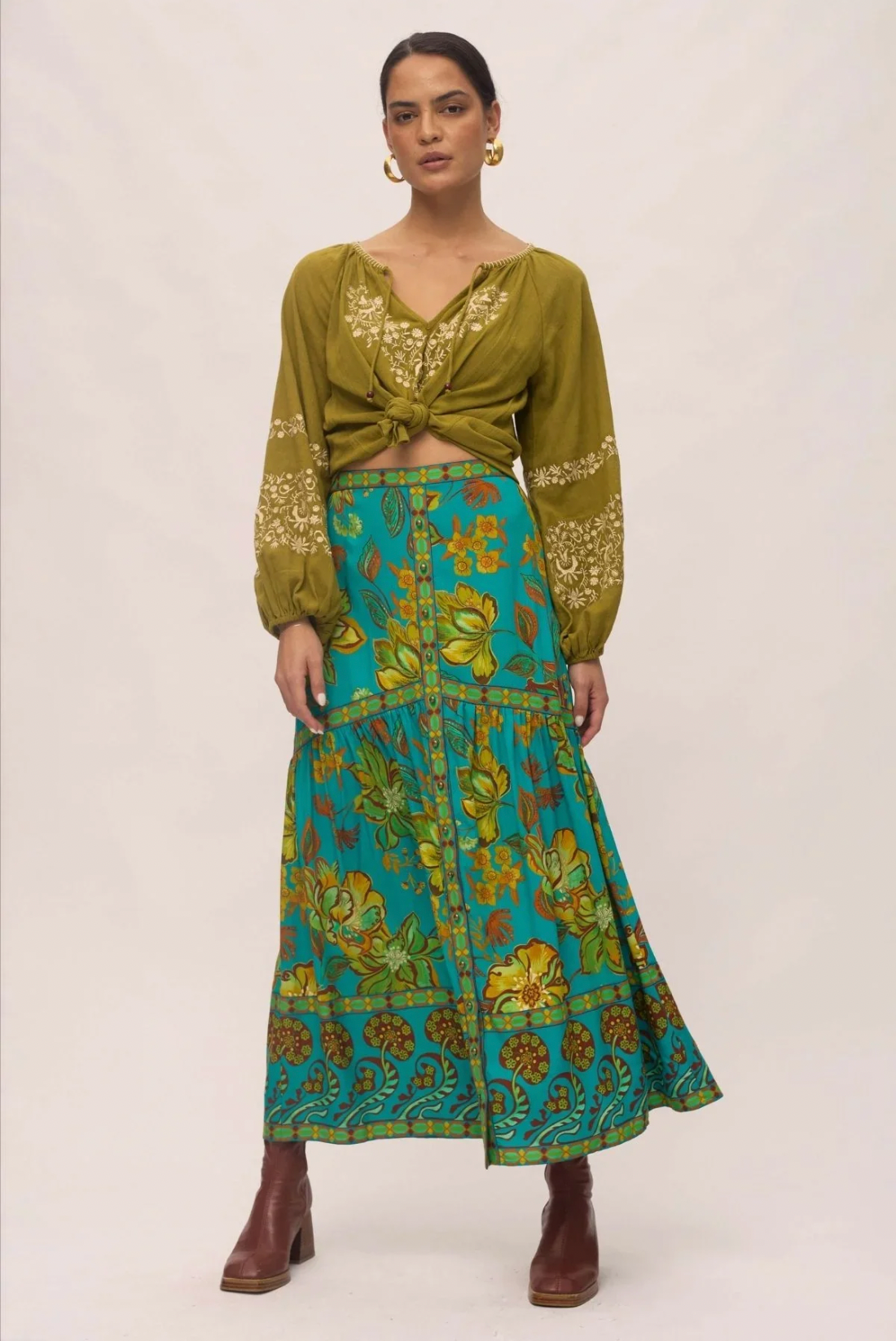 A woman wears the Nine Lives Bazaar Matilda Maxi Skirt in Peacock, styled with a green tie-front blouse, brown ankle boots, and gold hoop earrings. She stands against a plain background with her hair pulled back.