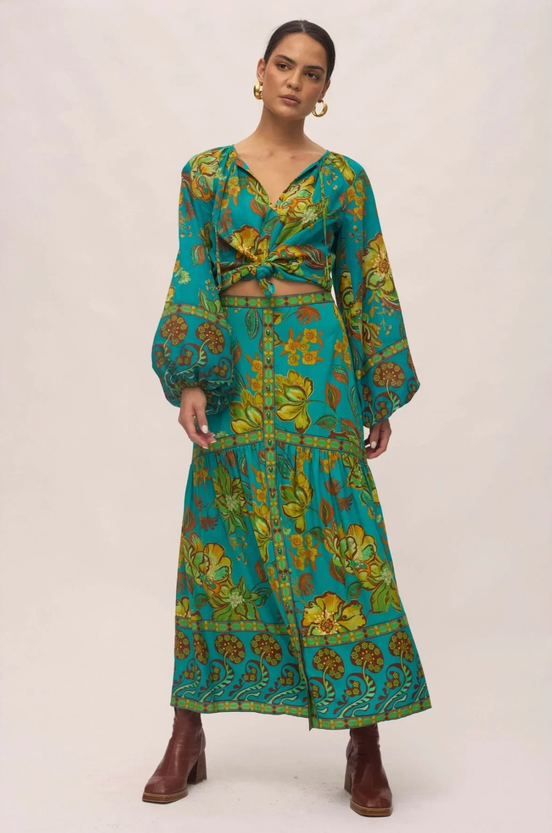 A woman models the Nine Lives Bazaar Matilda Maxi Skirt in Peacock, paired with a matching long-sleeve crop top. She accents her look with gold hoop earrings and brown ankle boots.