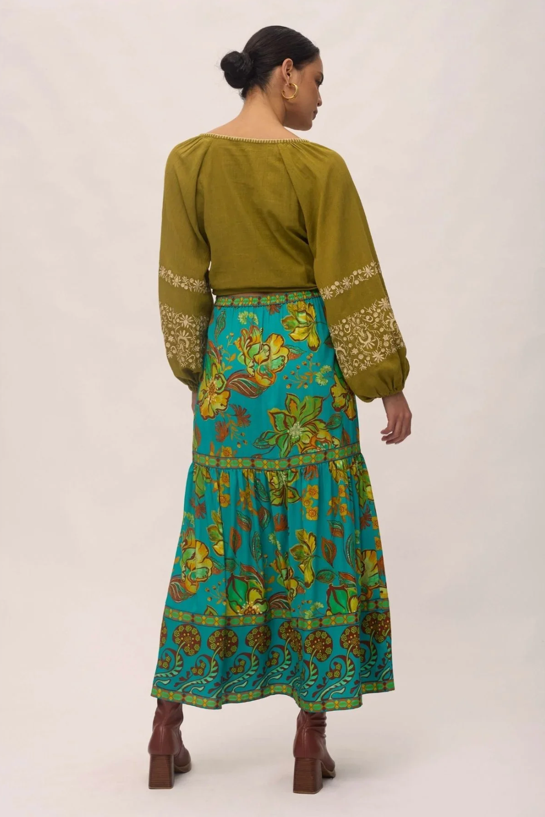 A woman with her dark hair in a low bun wears the Nine Lives Bazaar Matilda Maxi Skirt in Peacock, paired with an olive green embroidered long-sleeve blouse and brown boots, standing facing away.