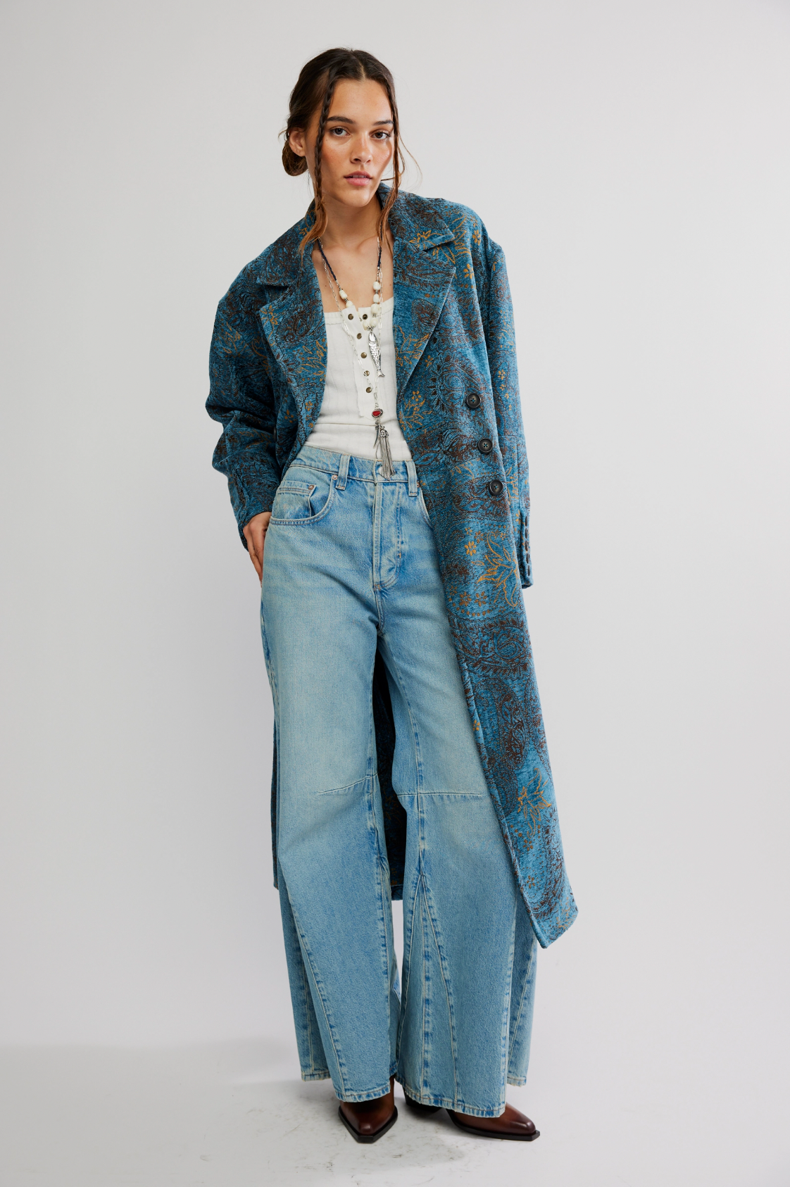 A woman wears the Micha Duster by Free People over a white top, light blue wide-leg jeans, and brown pointed shoes as she stands against a plain white background.