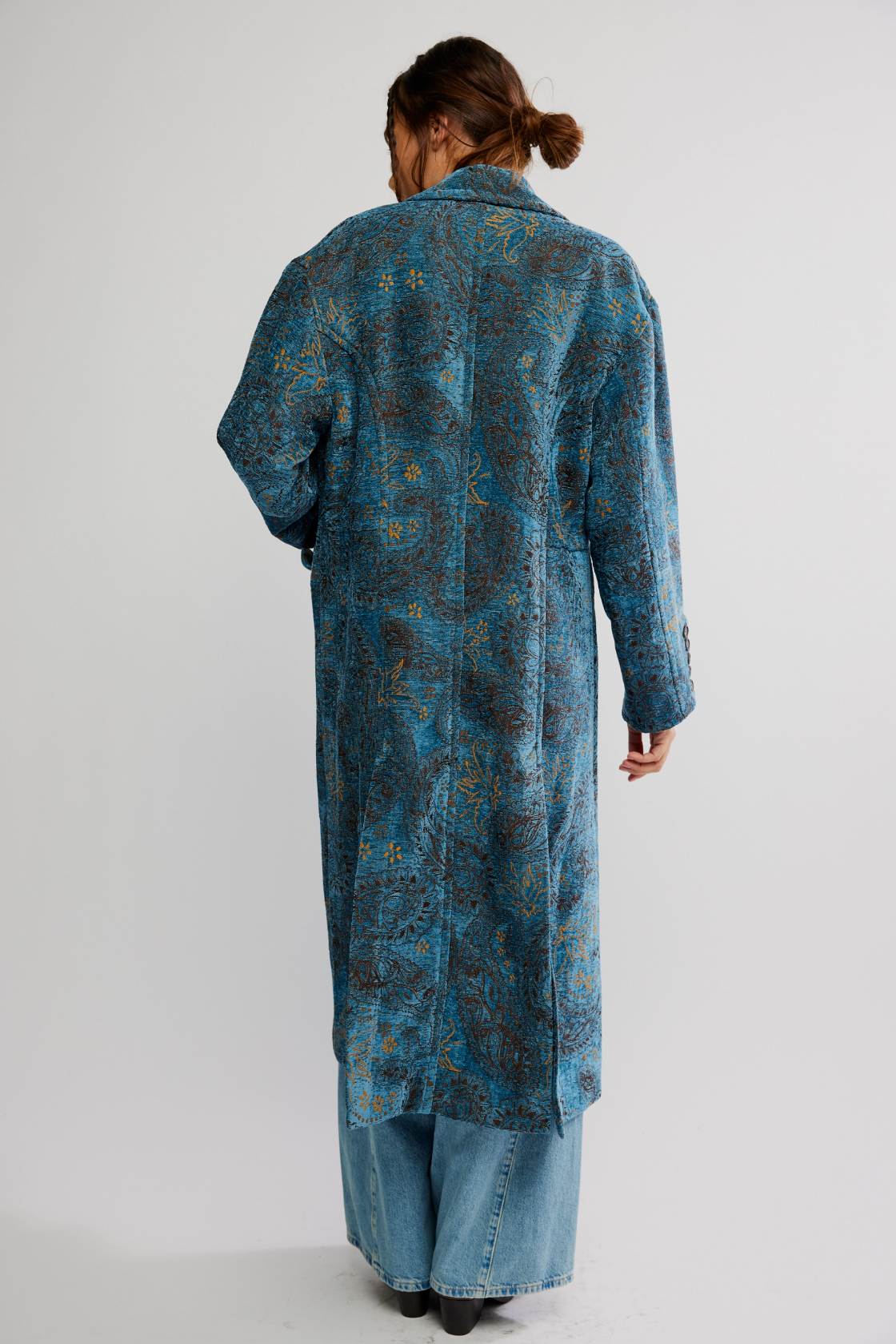 A person with brown hair in a low bun wears the Micha Duster by Free People, a longline blue patterned coat, and light blue wide-leg jeans, standing with their back to the camera against a plain white background.