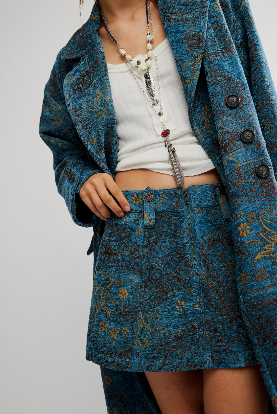 A person wears the teal and gold patterned Micha Duster longline coat by Free People with a matching skirt, a white ribbed crop top, and layered beaded tassel necklaces. Their face isn’t visible.