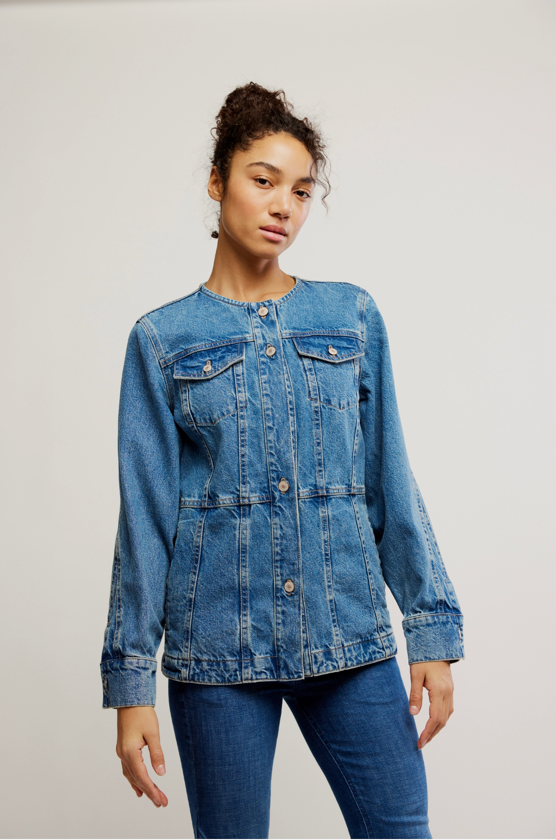 A woman with curly hair models the Lula Denim Jacket by Free People with matching jeans, standing against a light background and gazing confidently at the camera in a relaxed pose.