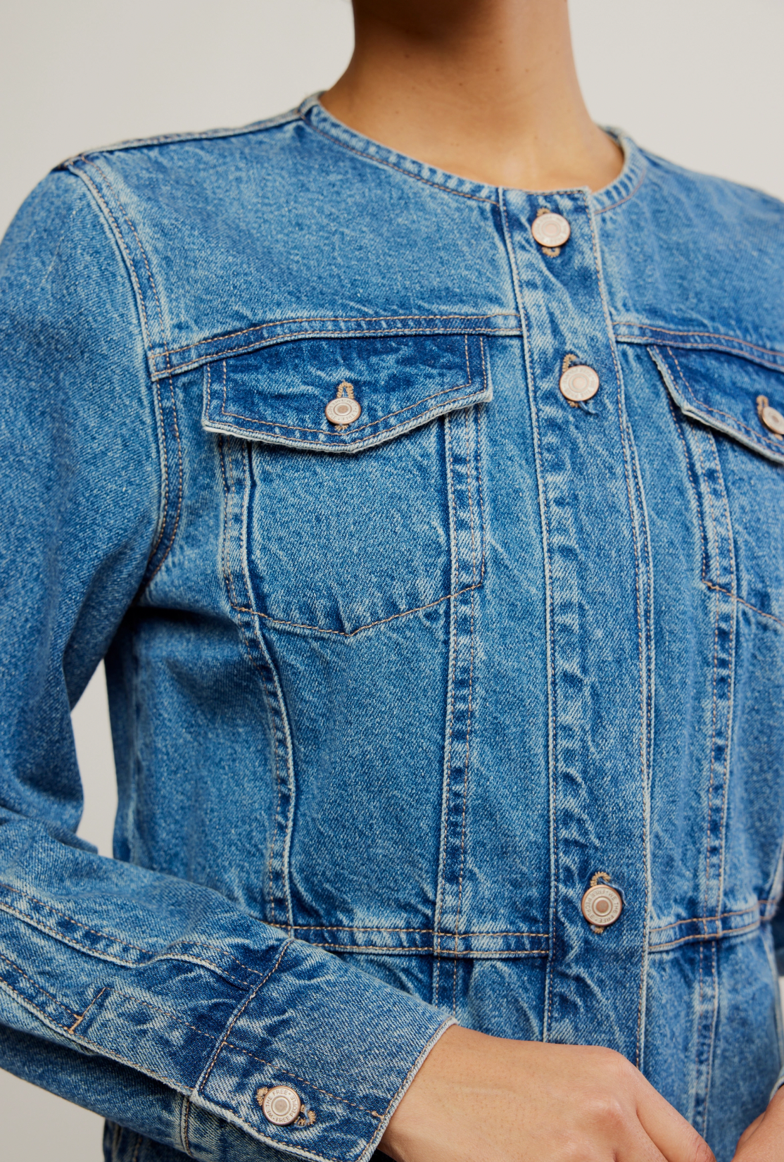 A person wears the Lula Denim Jacket by Free People, a blue collarless jacket with front pockets and metal buttons, visible from shoulders to waist. The womens layering jacket features a round neckline; their hand is near the buttons.