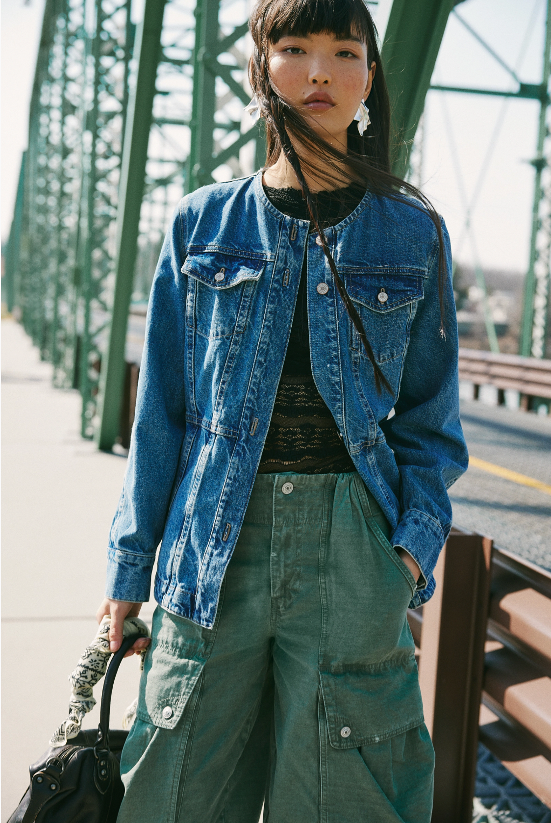 A woman stands on a bridge in the Lula Denim Jacket by Free People, layered over a black lace top and green cargo pants, with a black handbag. The green metal bridge complements her stylish look on this sunny day.