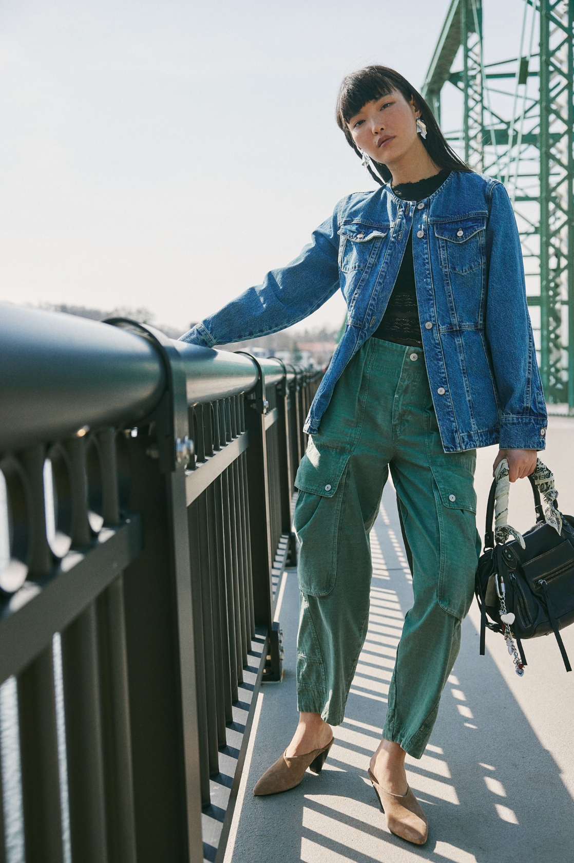 A woman stands on a bridge, leaning on the railing, wearing the Lula Denim Jacket by Free People, green cargo pants, brown shoes, and holding a black handbag. The green bridge structure and clear sky form the background.
