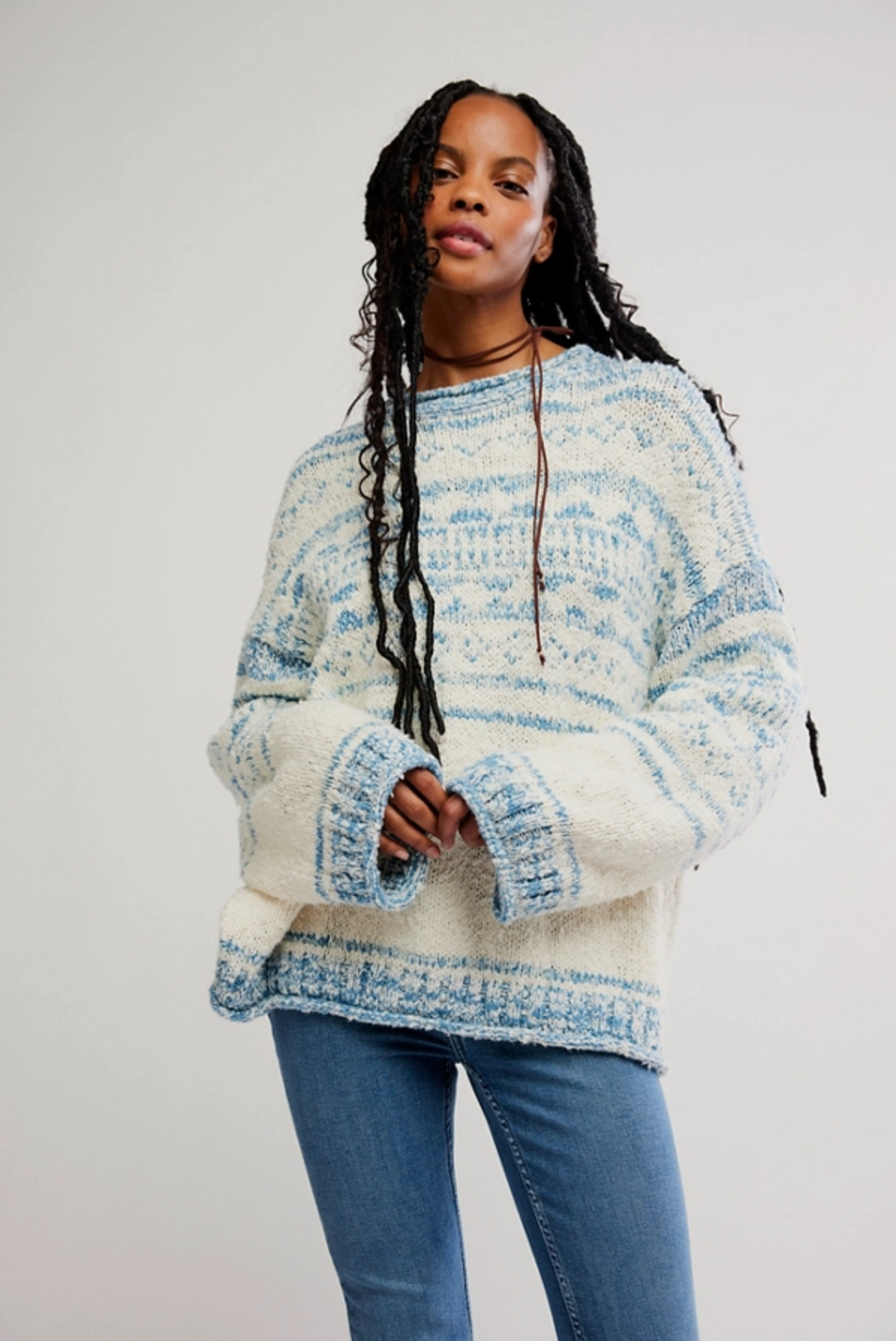 A person with long braids wears Free People’s oversized Dani Pattern Sweater and blue jeans, standing against a plain light background.