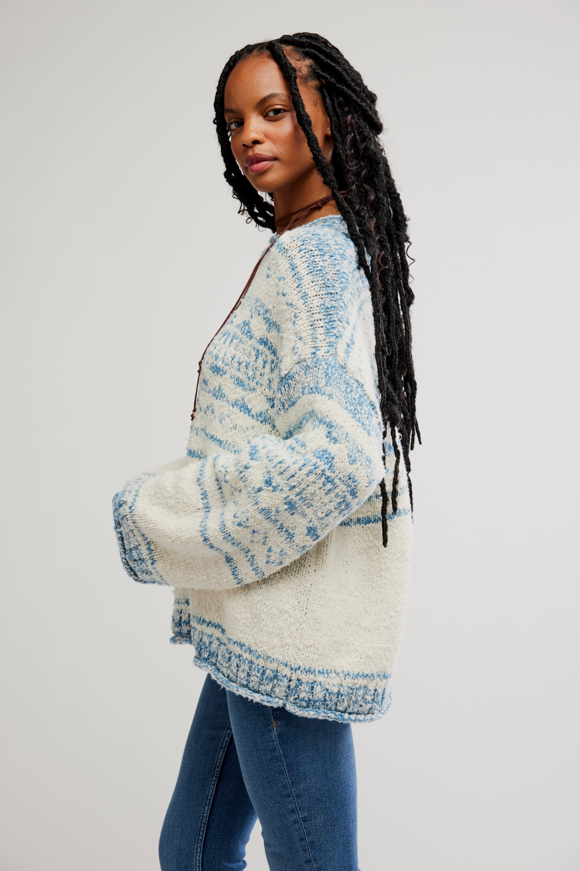 A woman with long braided hair wears the oversized Dani Pattern Sweater by Free People, featuring a blue and white Fair Isle pattern, with blue jeans, standing in profile against a plain light background.