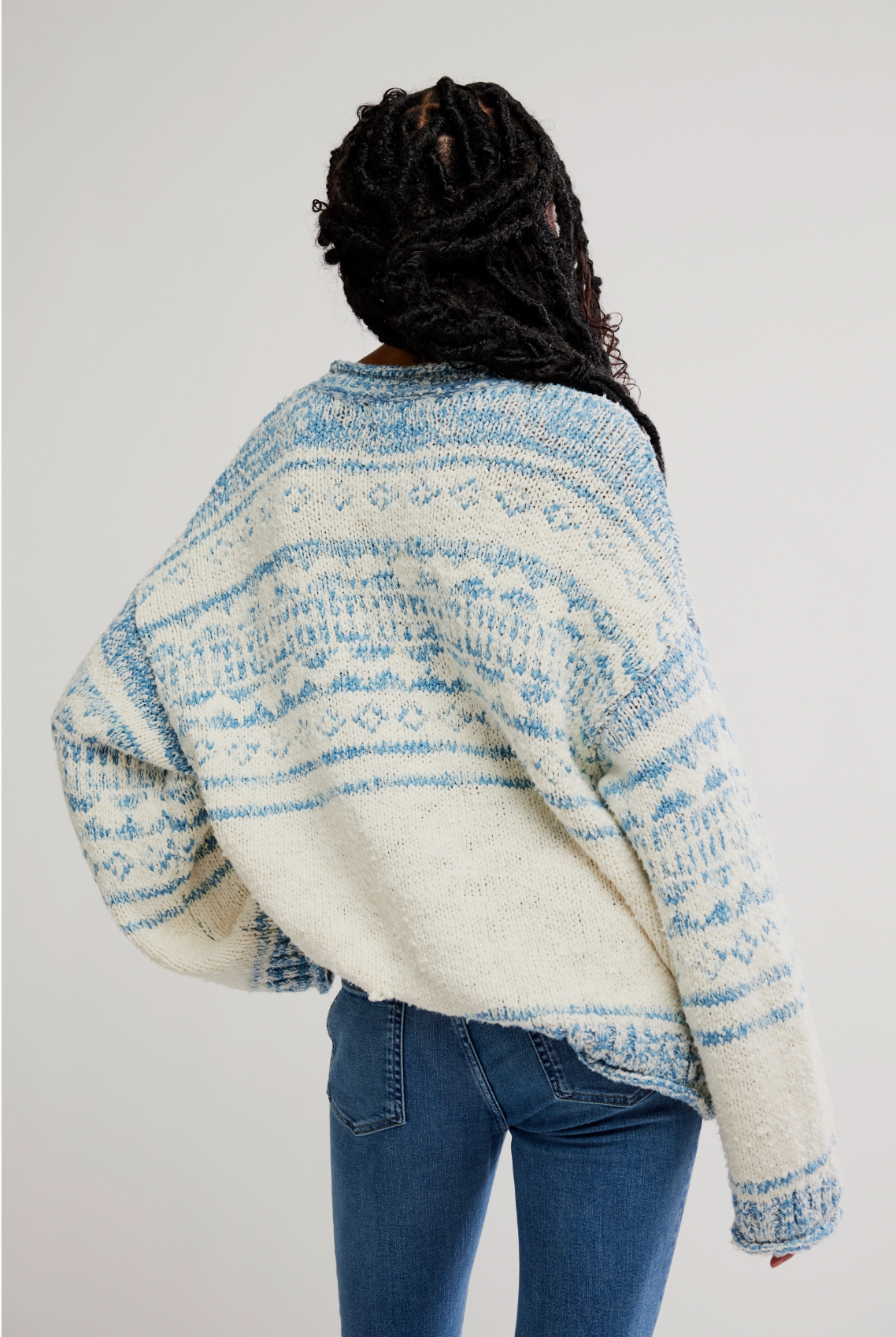 A person with long, dark braided hair is seen from behind wearing the Free People Dani Pattern Sweater in white and blue with blue jeans, standing against a plain light background.