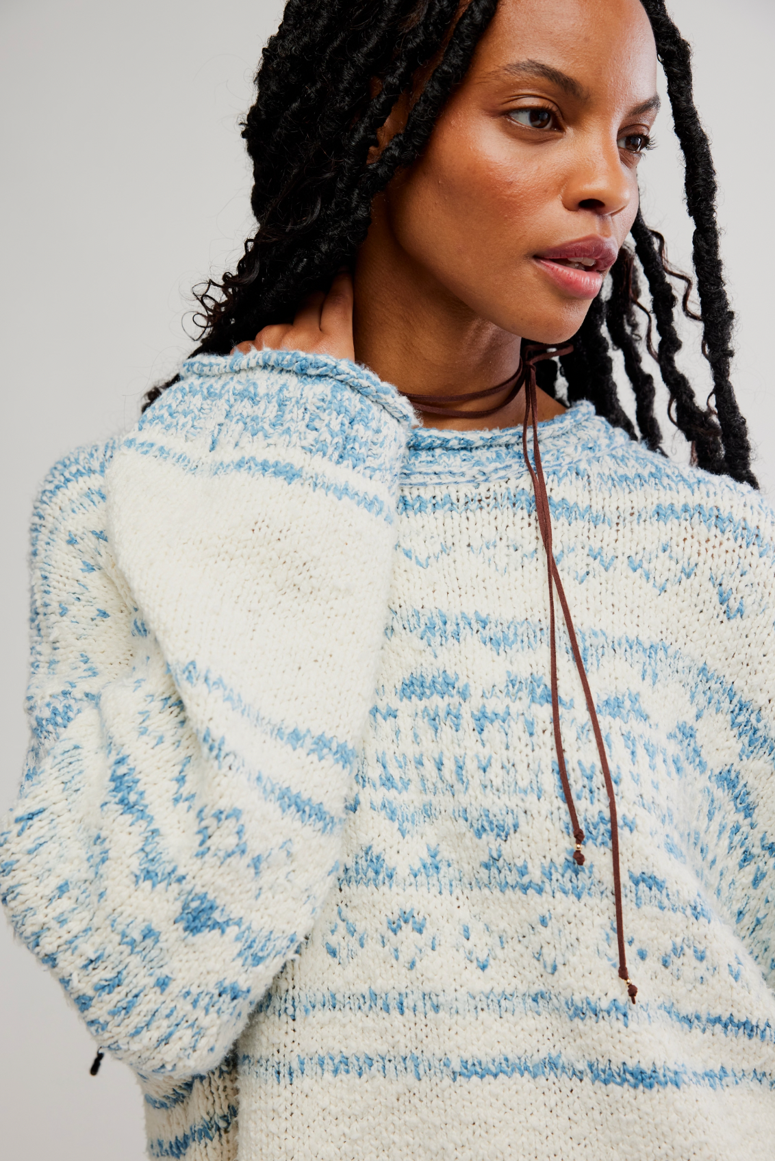 A person with long, dark braids wears the oversized Dani Pattern Sweater by Free People in white and light blue, looking to the side with one hand on their neck against a plain, light-colored background.