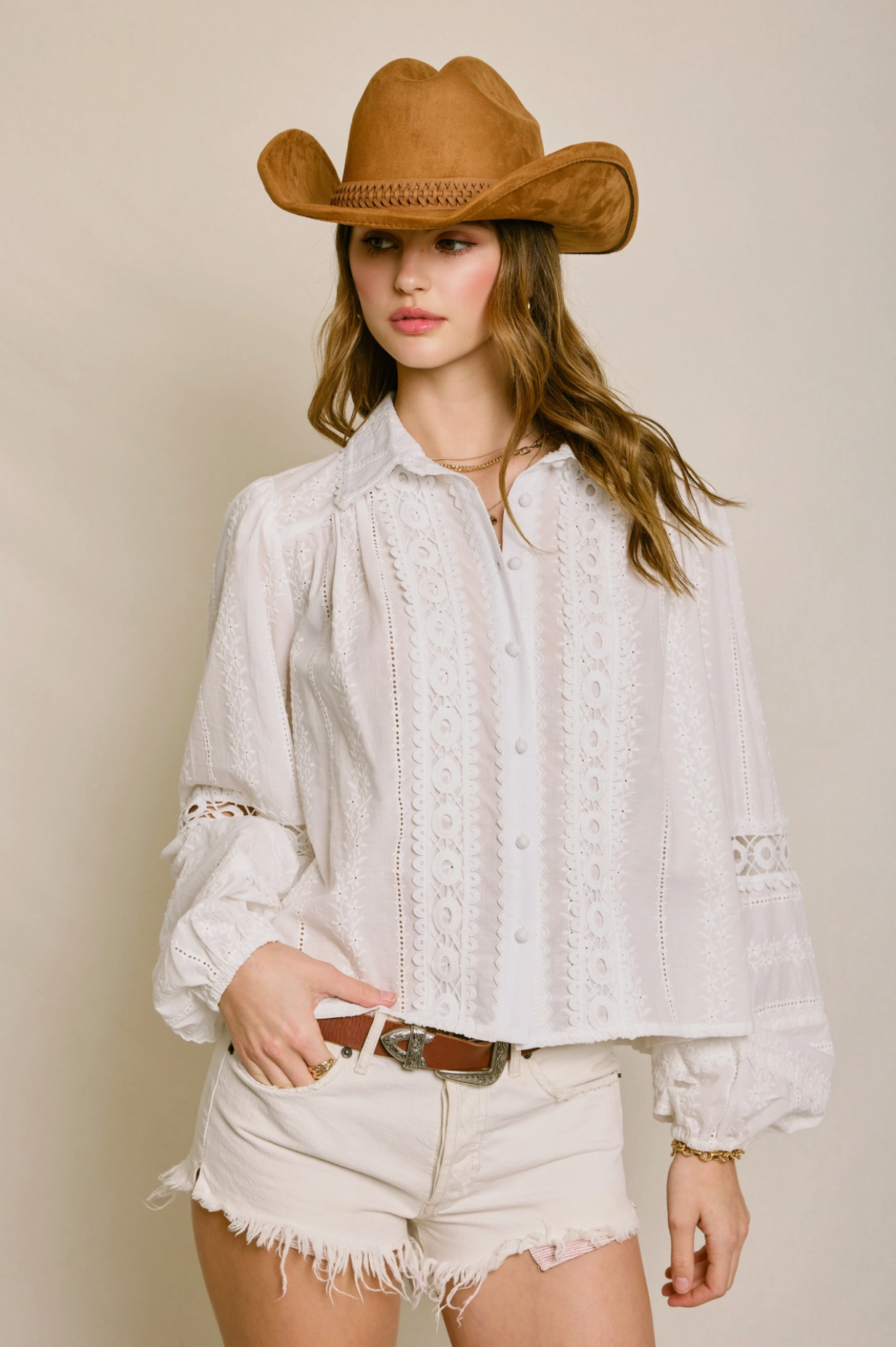 A woman poses against a neutral background wearing the Dress Forum Sunbeam Eyelet Blouse, a brown cowboy hat, and white frayed shorts, with one hand on her belt.