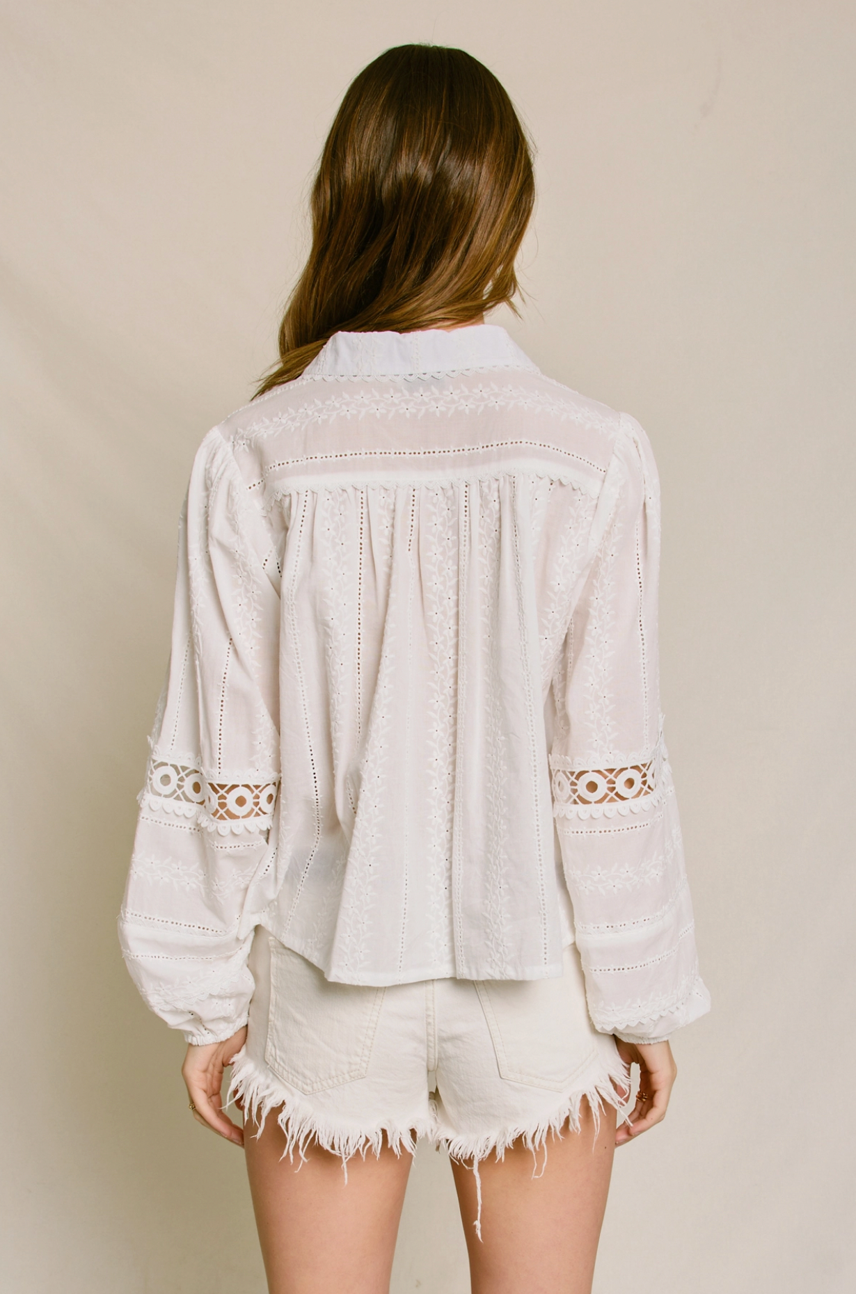 A woman with long, wavy brown hair stands with her back to the camera, wearing the Dress Forum Sunbeam Eyelet Blouse and white frayed-edge shorts against a plain, neutral background.