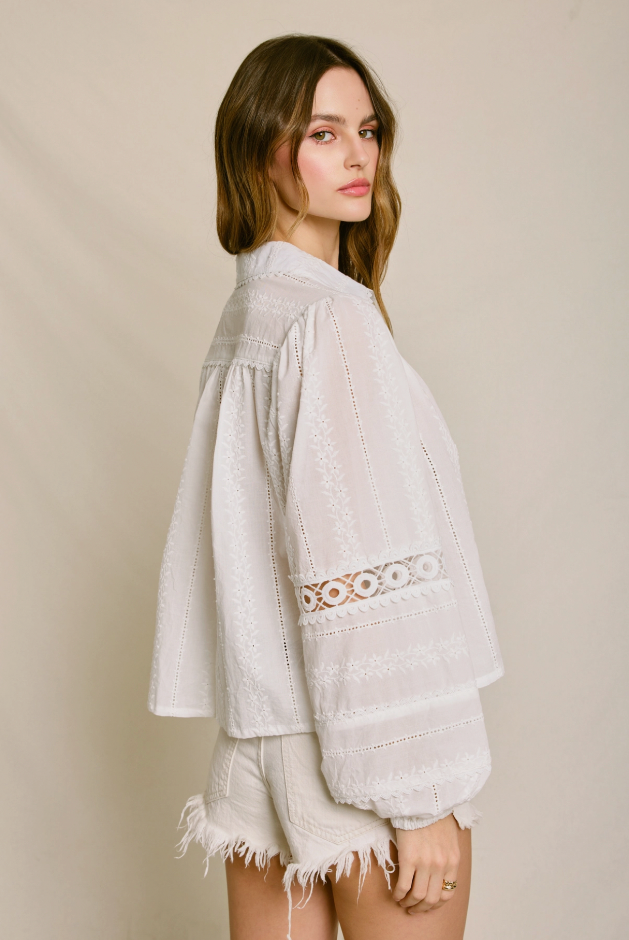 A woman with long brown hair stands sideways, looking at the camera. She wears the Sunbeam Eyelet Blouse by Dress Forum with wide sleeves and white frayed shorts against a neutral background.