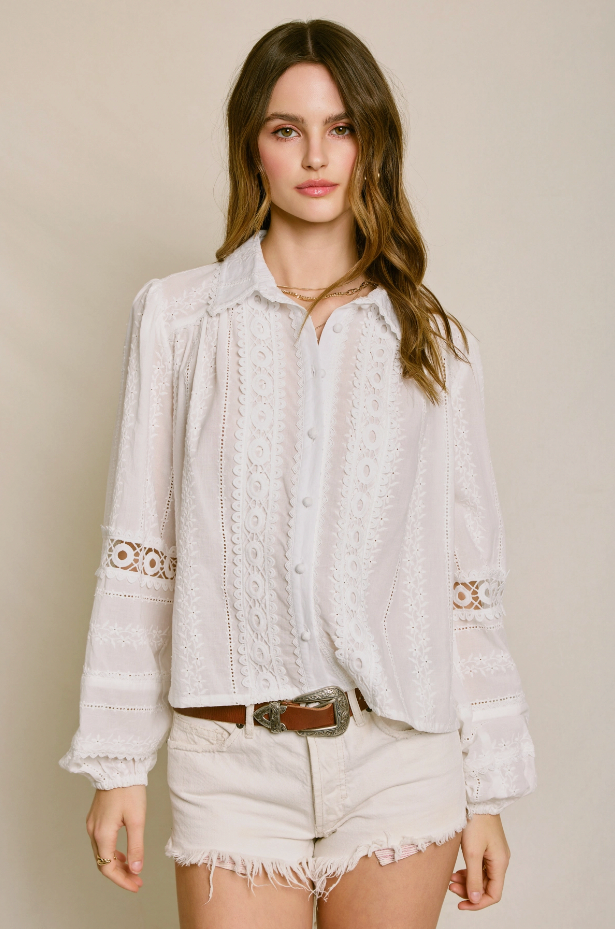 A woman with wavy brown hair wears the Dress Forum Sunbeam Eyelet Blouse and cream frayed shorts, standing against a beige background with a neutral expression and her left hand at her side.